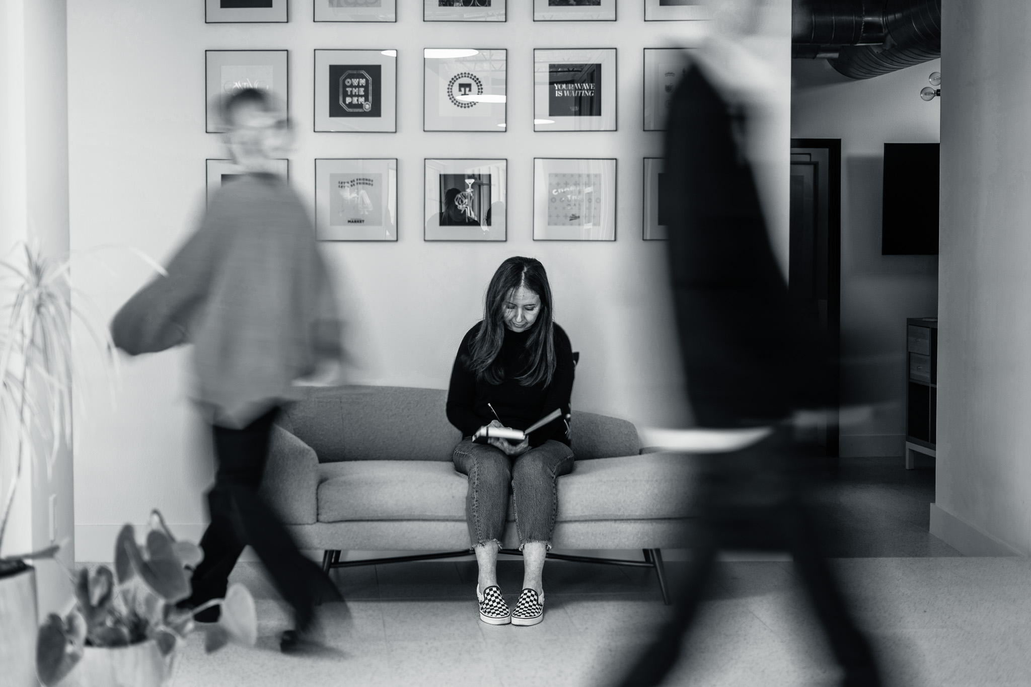 A woman sits on a sofa reading a book in a modern room with framed pictures on the wall, while two people walk past in the foreground, blurred from motion.
