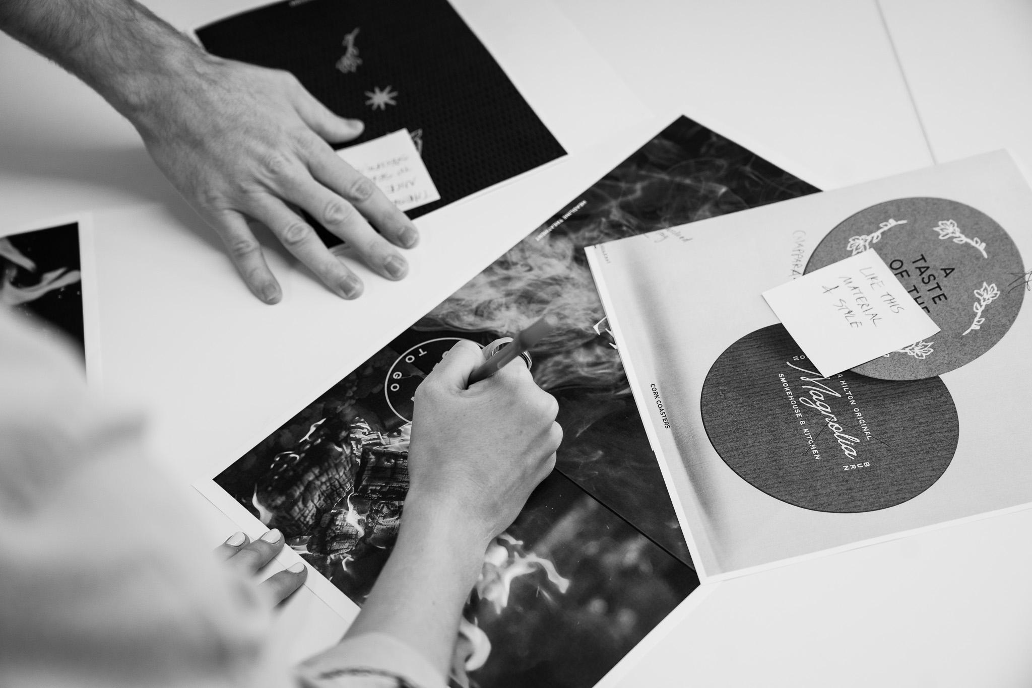 Two people work on printed design materials at a table, with hands visible arranging and pointing at various sheets and notes.