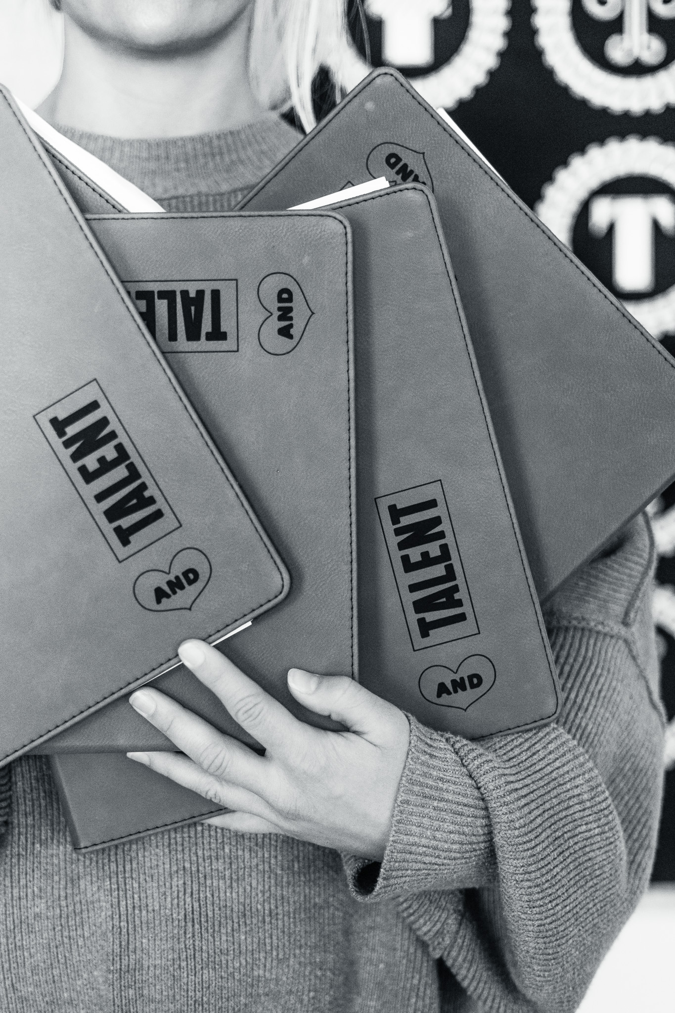 A person in a sweater holds three notebooks labeled "TALENT AND" in large black letters, partially covering their face.