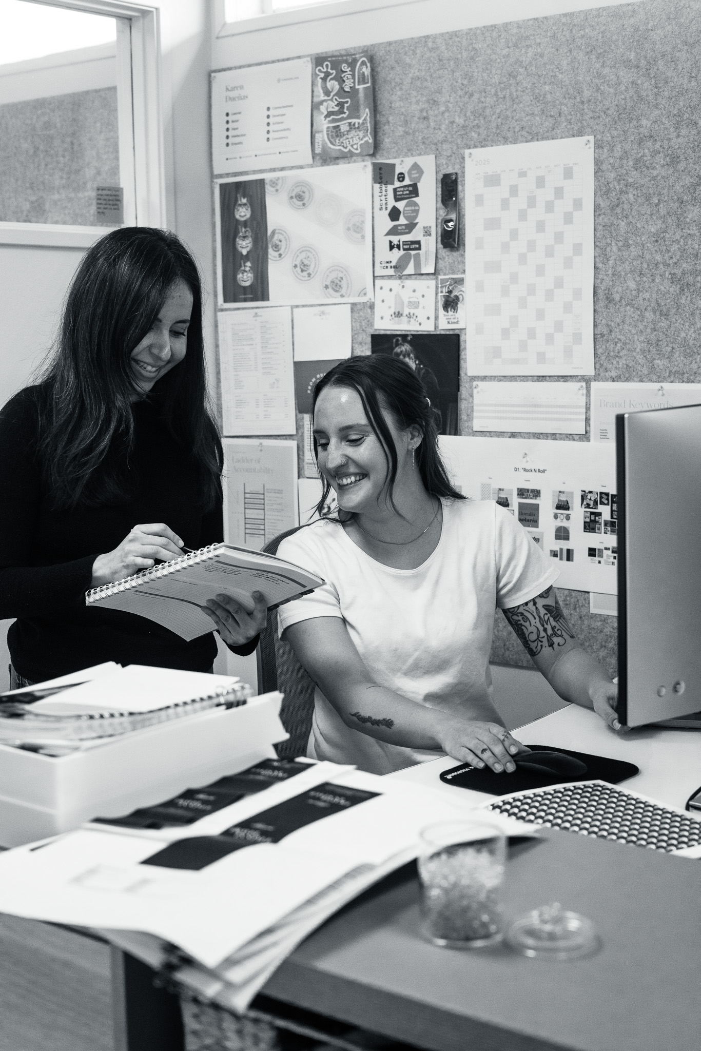 Two women in an office look at a notebook together, one seated at a desk with a computer mouse, the other standing and writing, surrounded by papers and design materials.