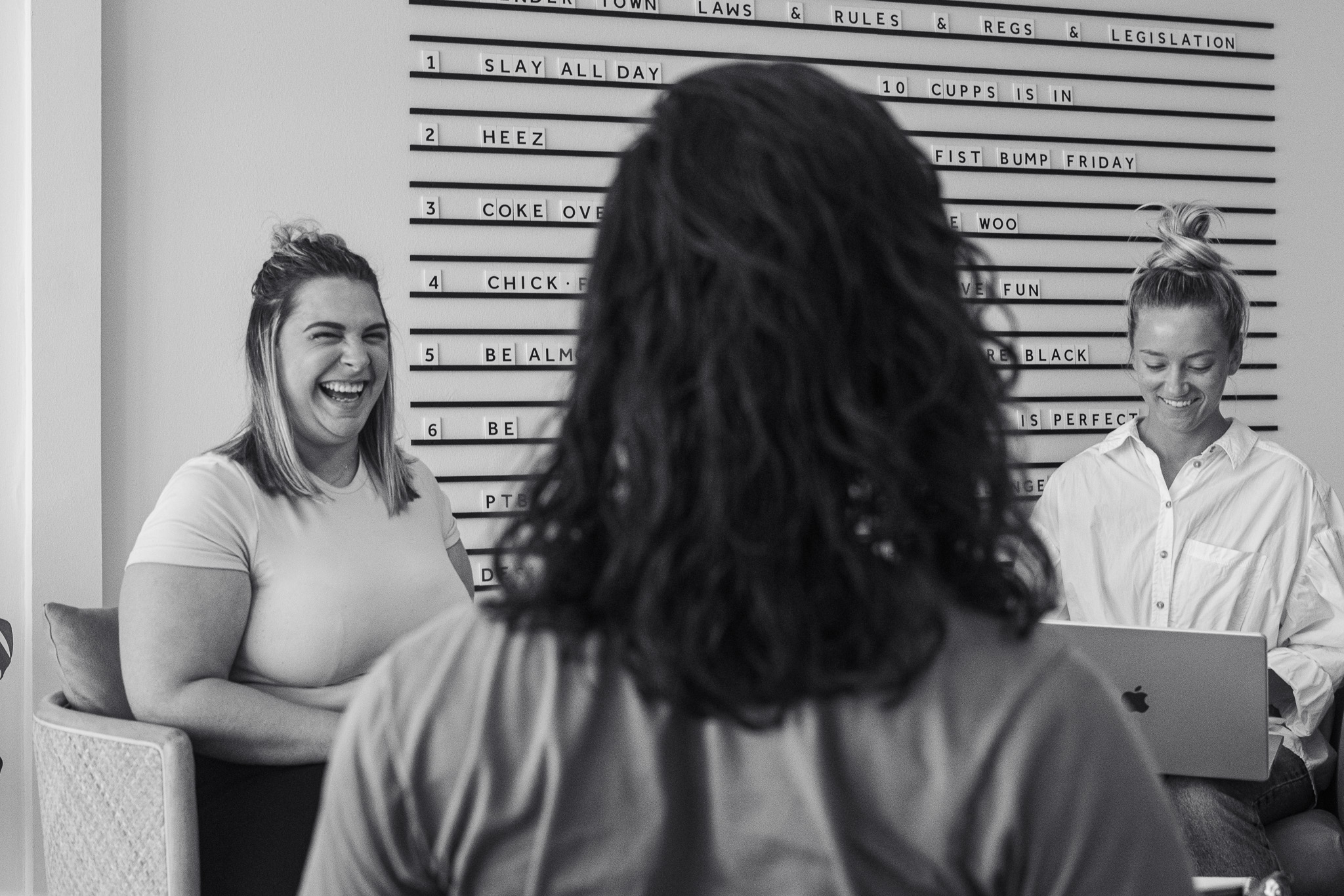 Three people are in an office; one woman is laughing, another is typing on a laptop, and a third person is seated with their back to the camera.