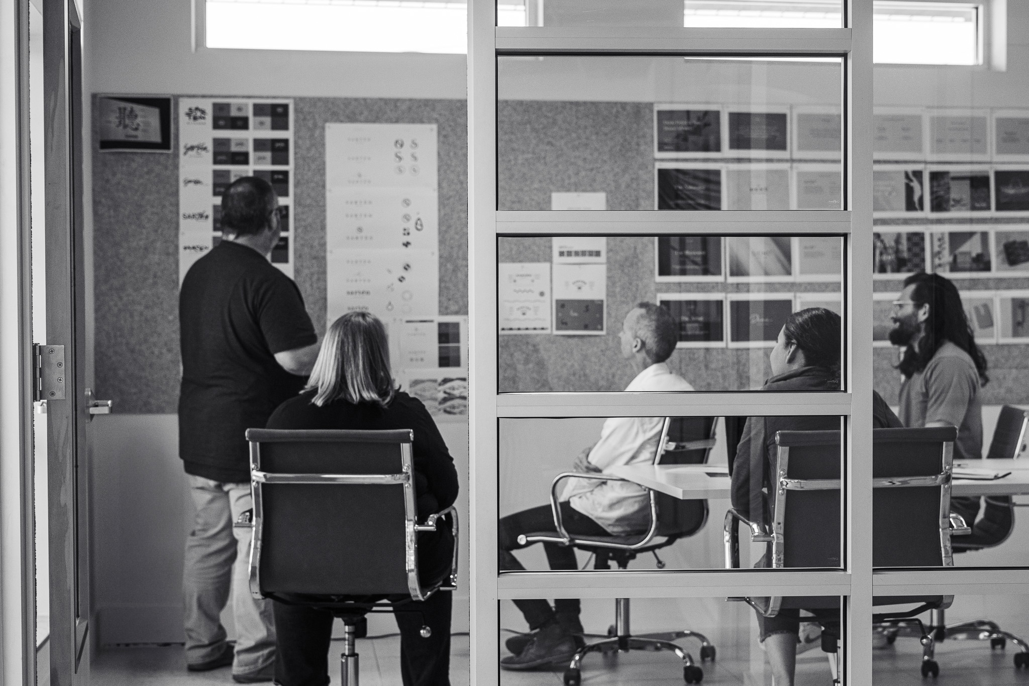 A group of people sits at a conference table while one person stands and presents materials on a board in a modern meeting room.