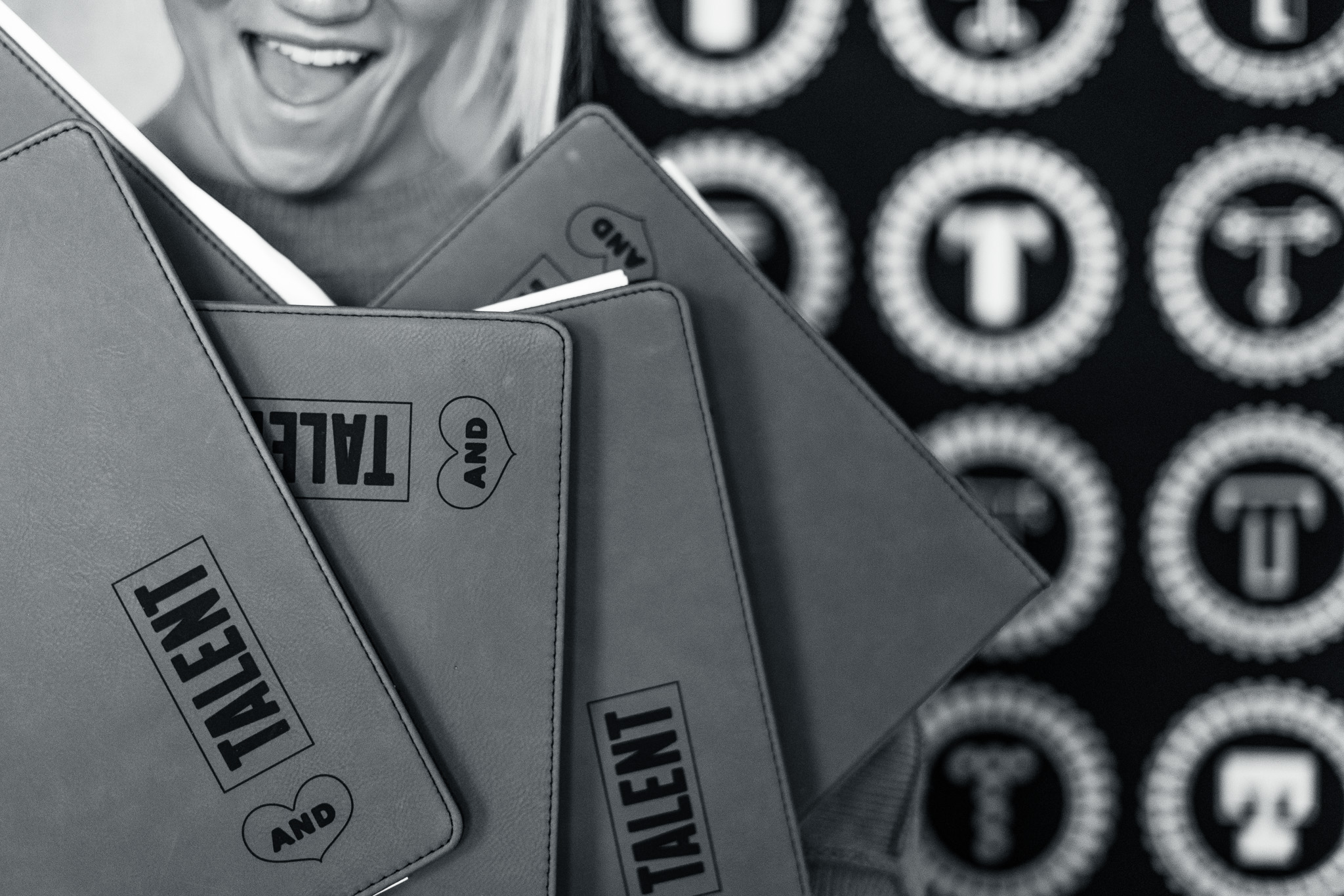 Black and white photo of several notebooks labeled "TALENT AND" stacked together, with part of a smiling person's face and a patterned background featuring the letter "T.