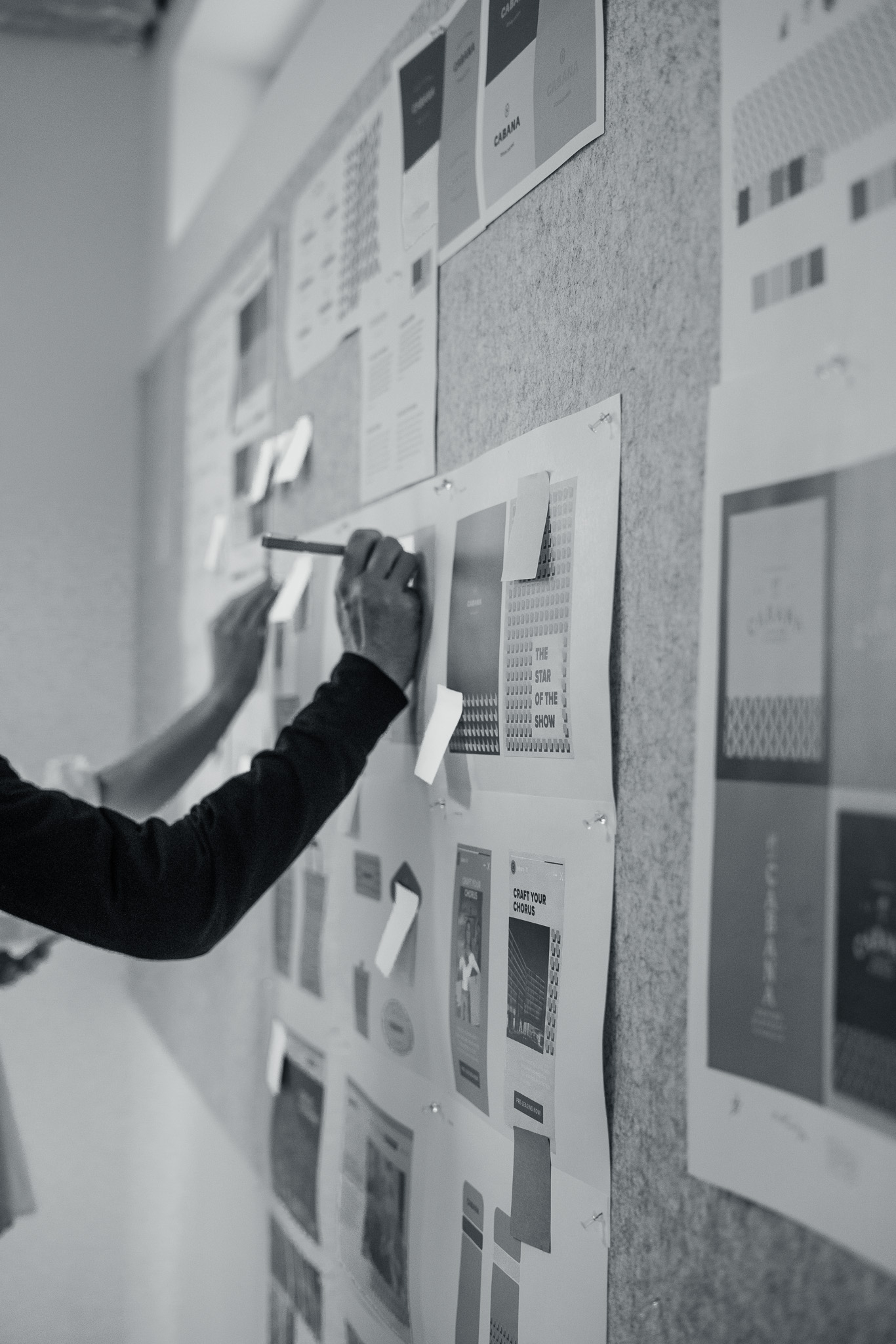 A person uses a ruler to measure or align papers with design layouts pinned to a wall or board.