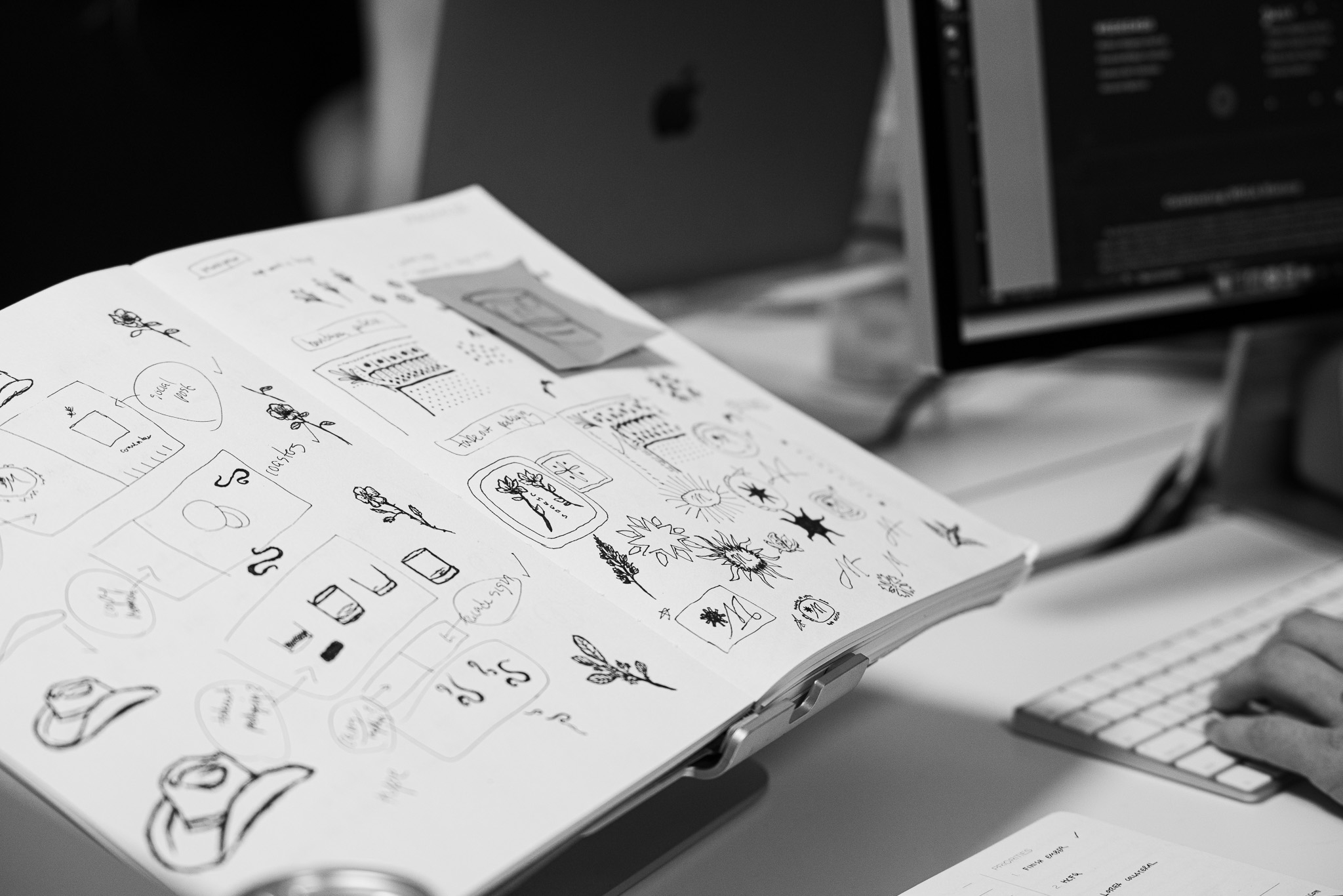 Open sketchbook with hand-drawn diagrams and doodles on a desk next to a computer monitor, keyboard, and a laptop in the background.