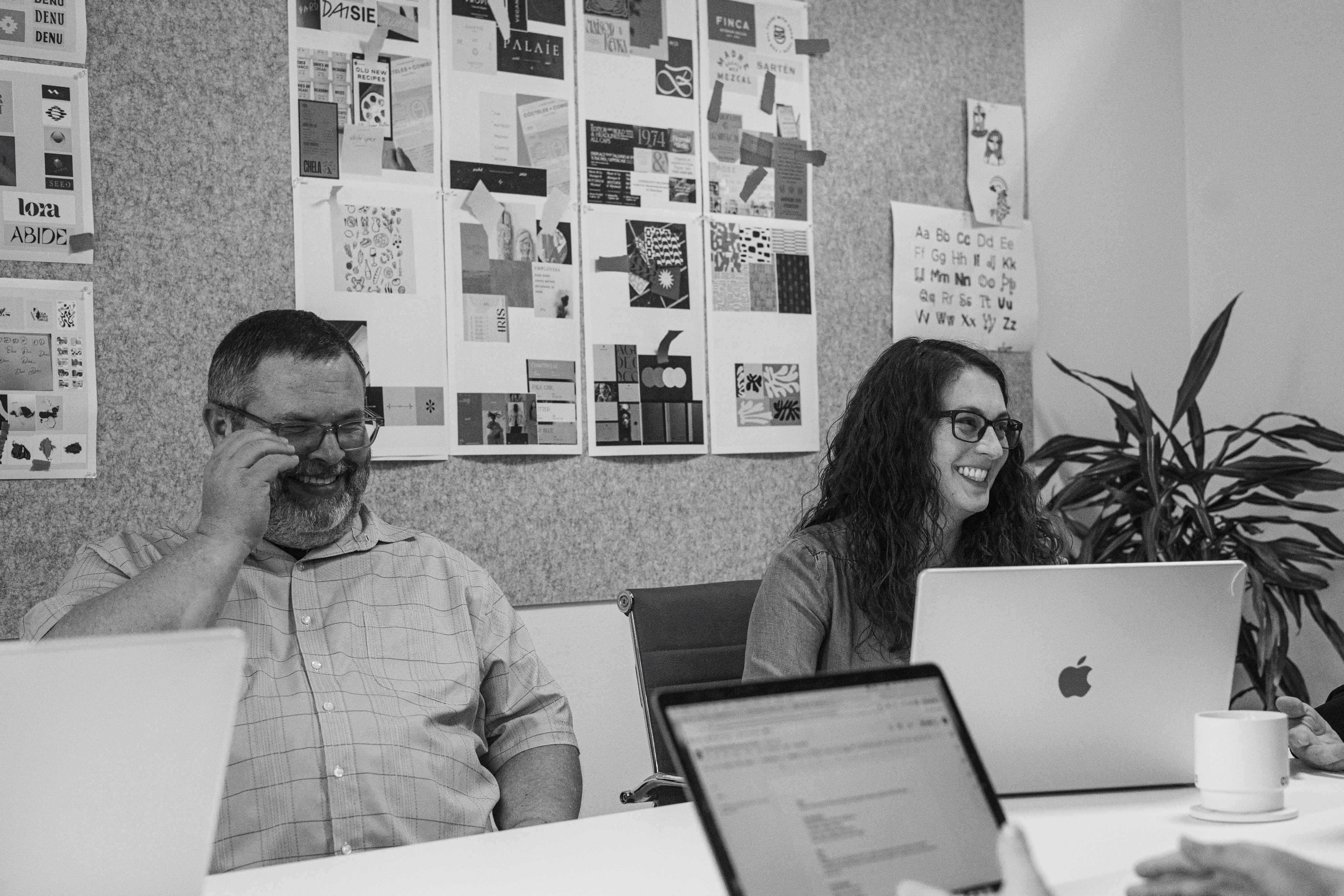 Two people sit at a conference table with laptops, smiling, in a meeting room decorated with various design posters on the wall.