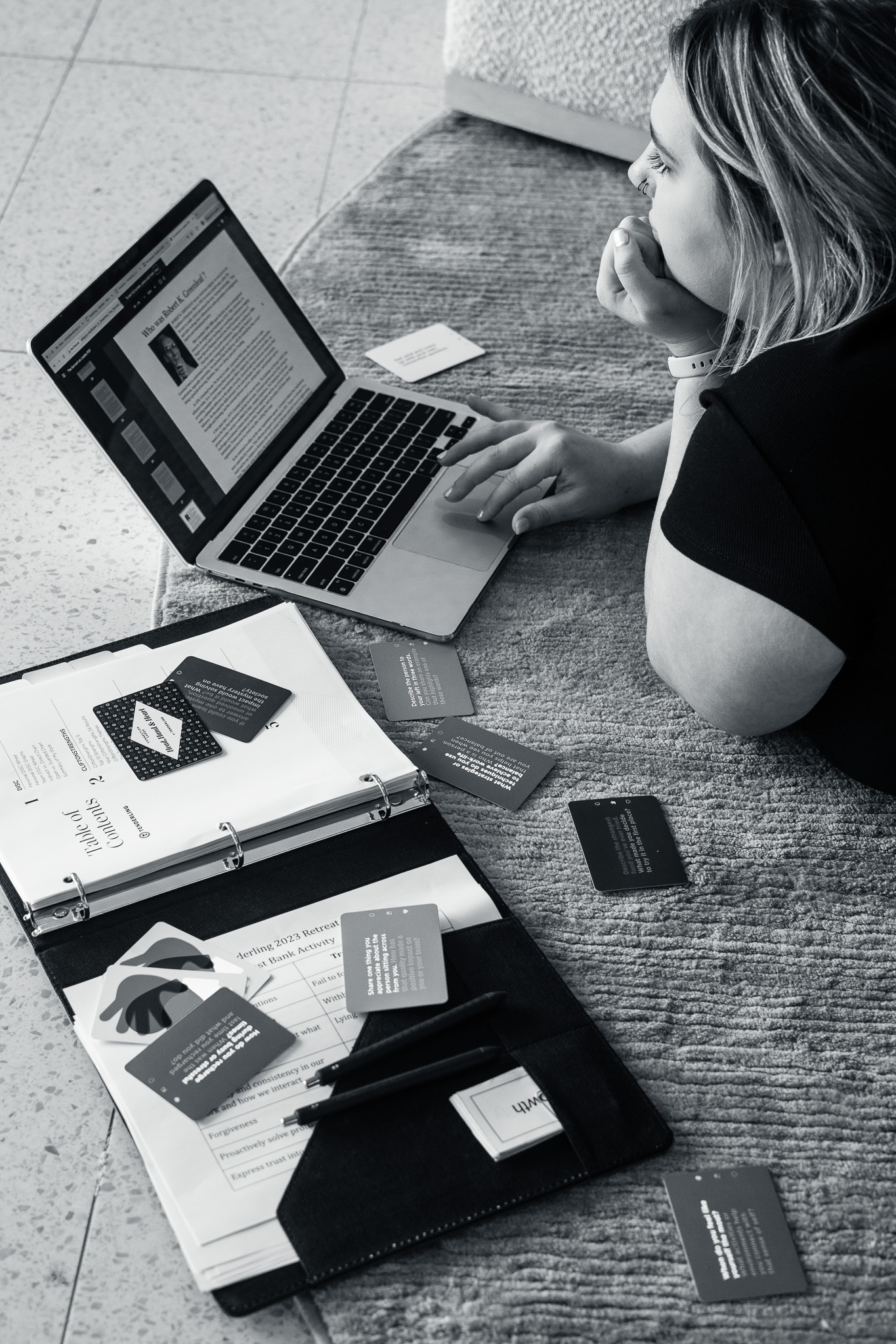 A person lies on the floor using a laptop, surrounded by scattered cards and an open binder with documents and pens.