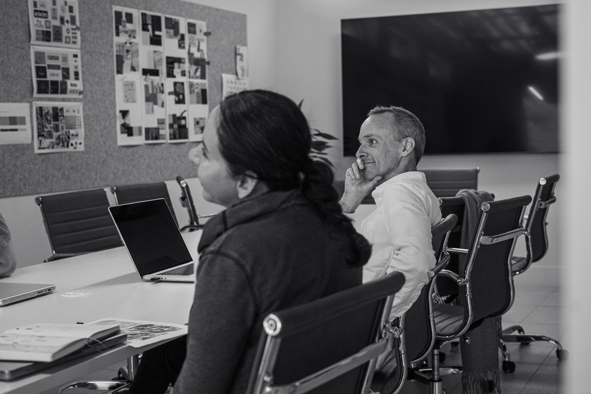 Two people sit at a conference table in a meeting room, one with a laptop open, with a large display screen and photos on the wall in the background.