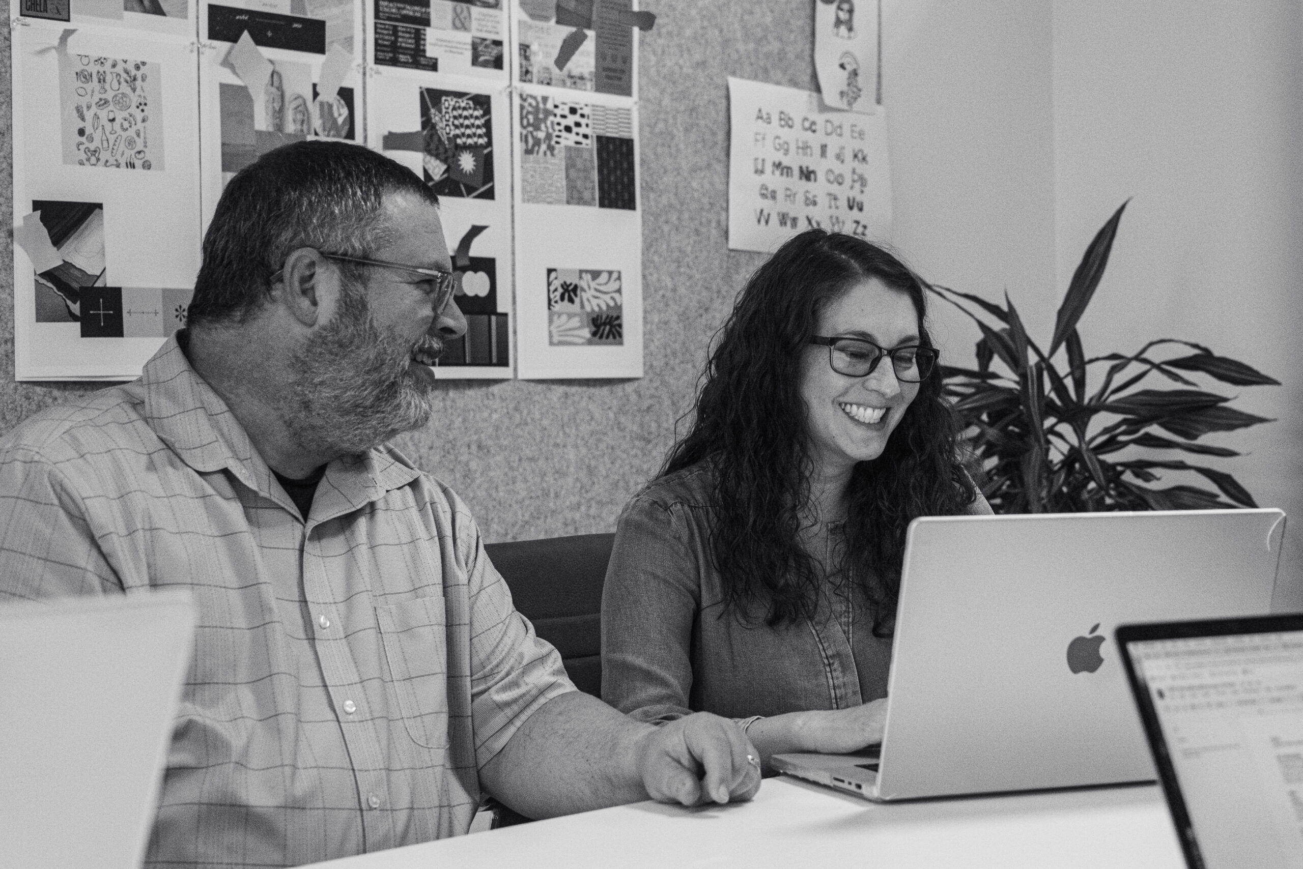 Two people sit at a table, smiling and working on a laptop, with design materials and a plant in the background.