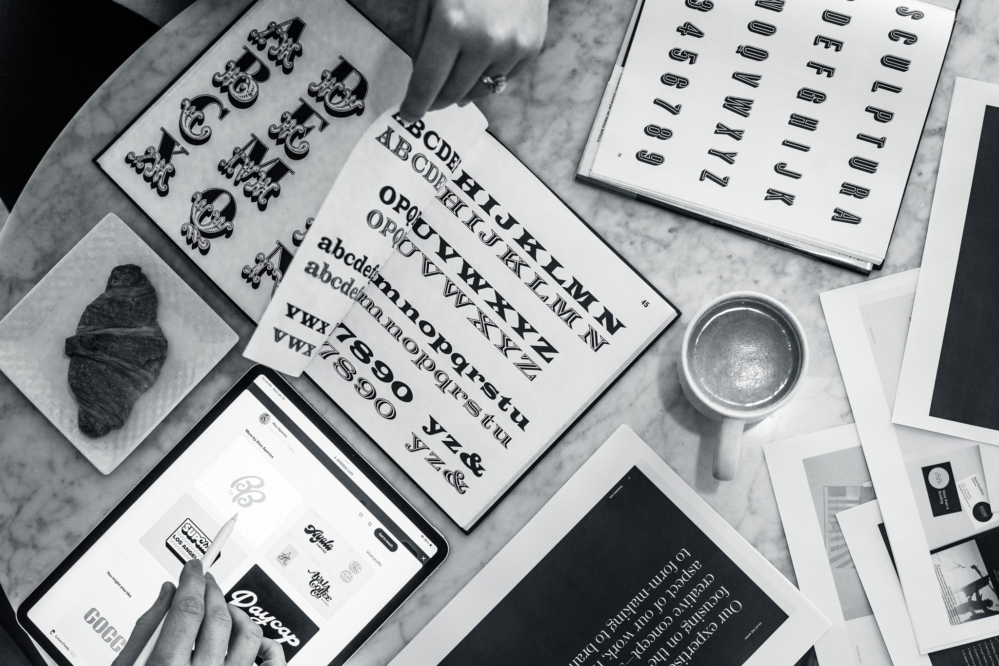 A tabletop with hands browsing various typefaces and fonts on paper and a tablet, alongside a cup of coffee and a croissant on a napkin.