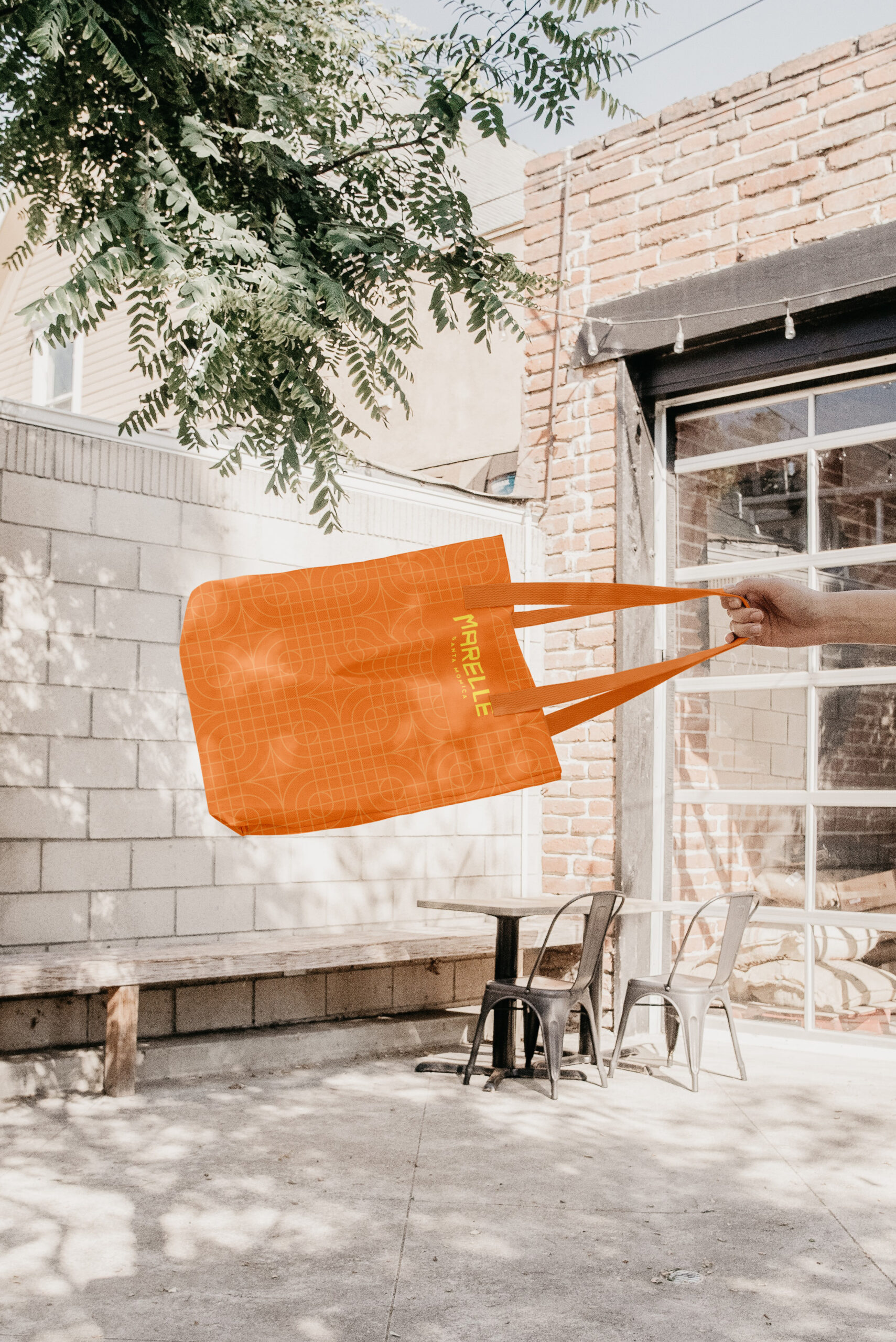 A hand holds an orange tote bag with "FRAGILE" written on it, outdoors near a brick building, metal chairs, and a concrete wall.