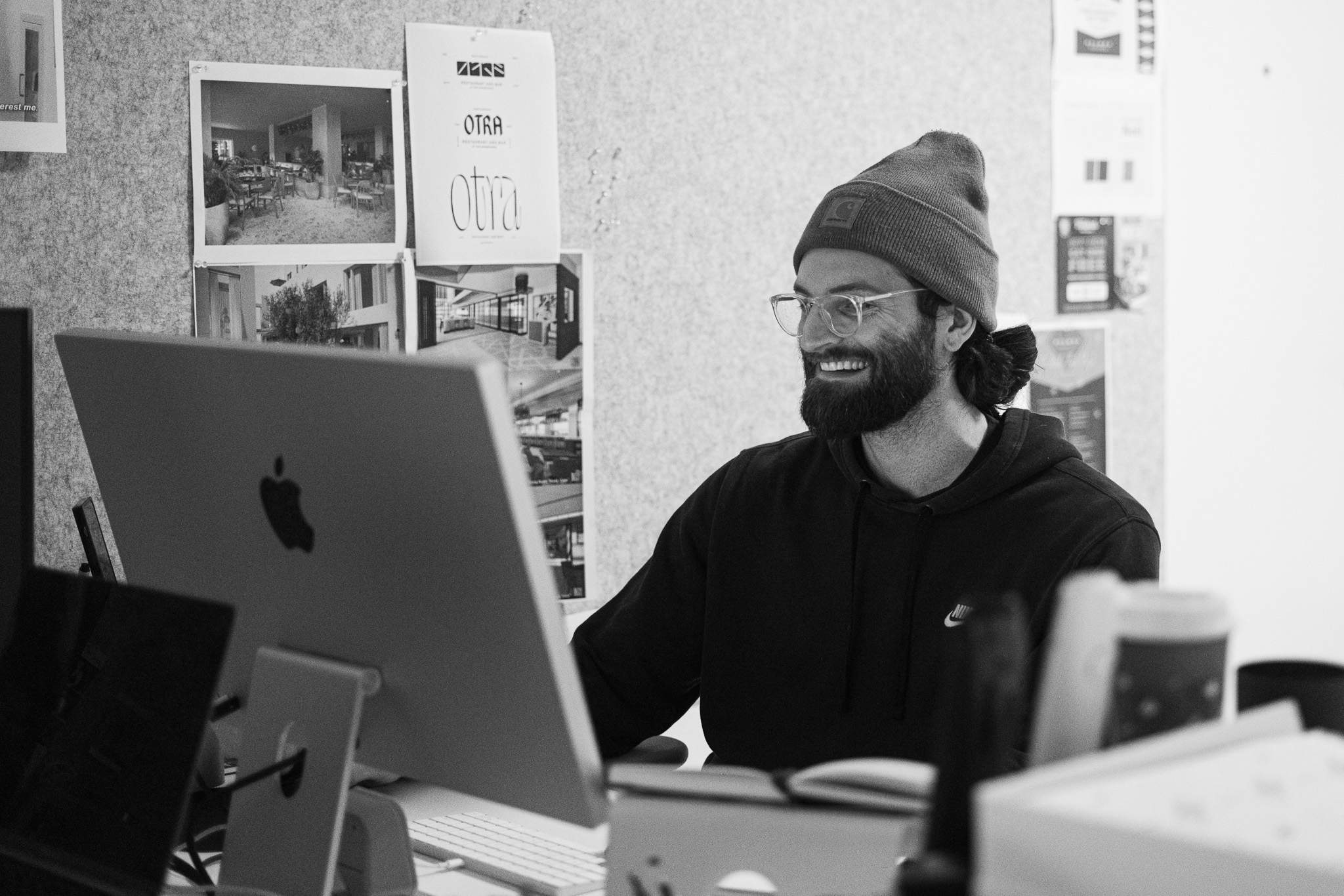 A man wearing glasses and a beanie sits at a desk, smiling while working on a large desktop computer in an office filled with design posters.