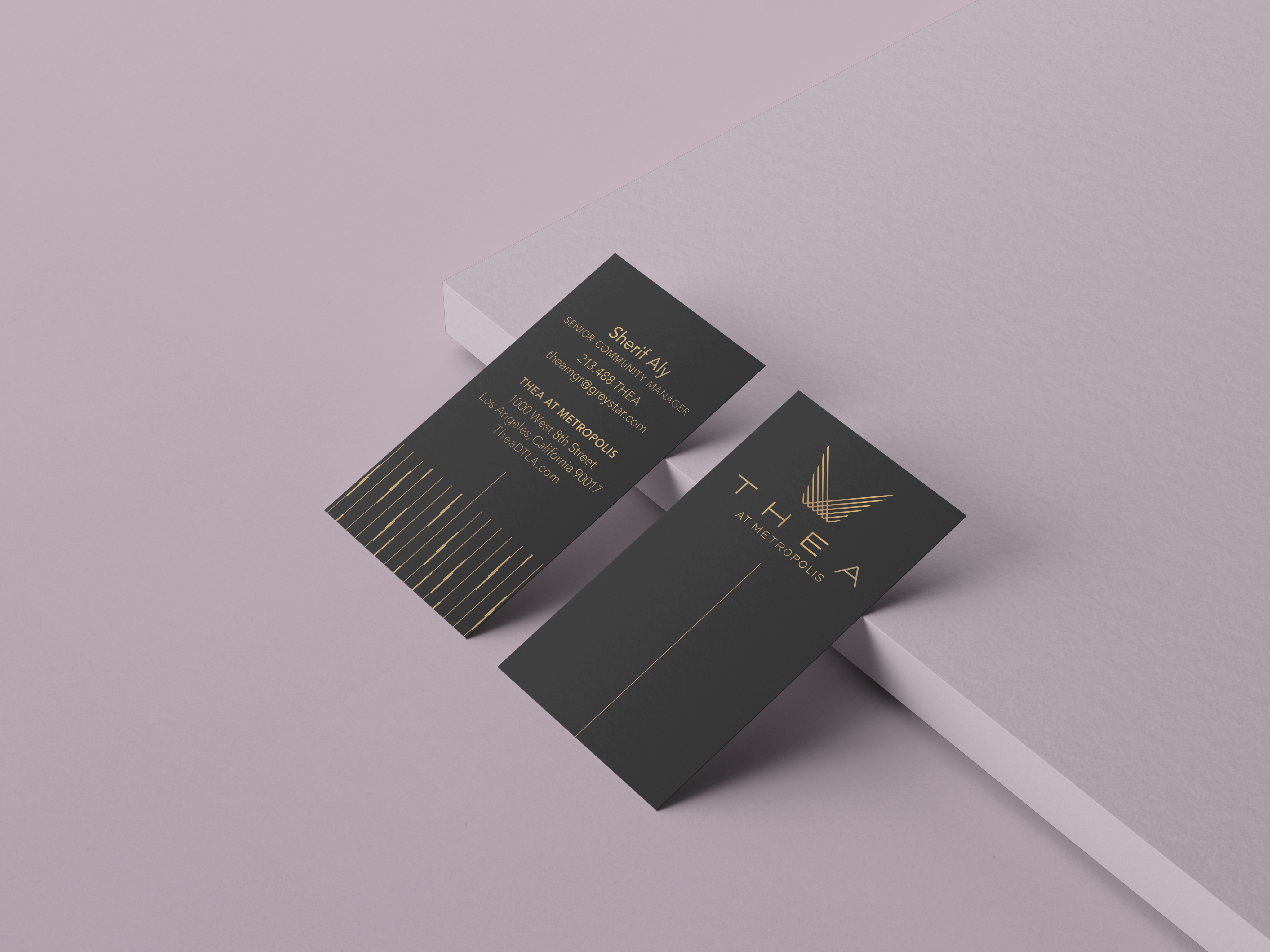 Two black business cards with gold text and logo are displayed on a light pink surface, one card facing up and the other showing contact details.