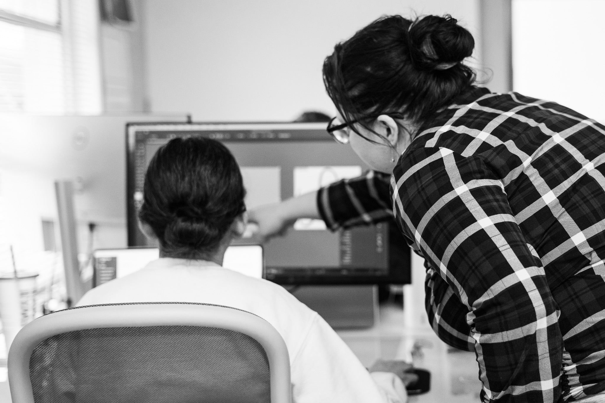 Two people work at a computer; one seated faces the screen, while the other stands and points at something on the monitor.