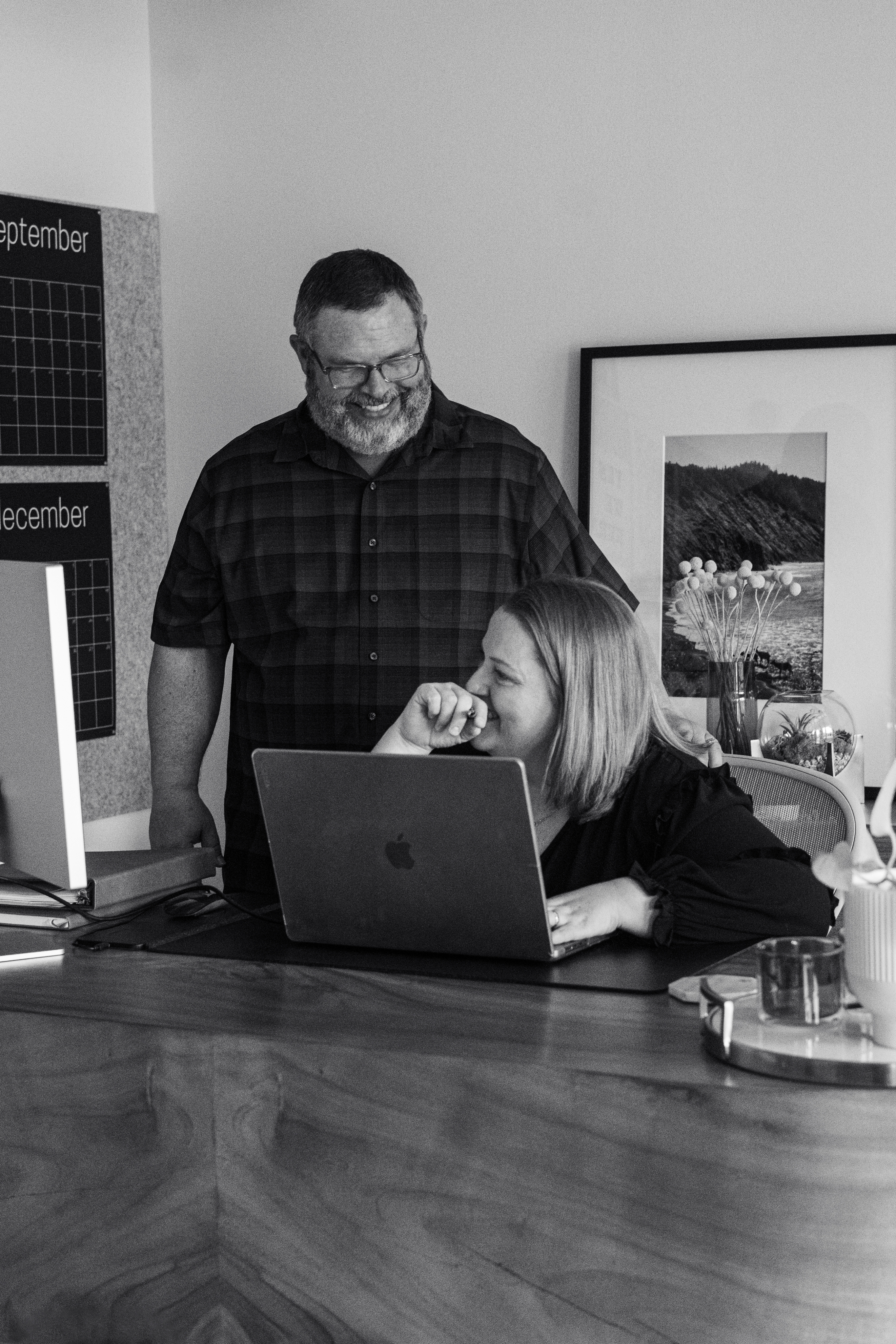 A man stands next to a woman seated at a desk with a laptop; they are both smiling. A calendar, framed prints, and office items are visible in the background.