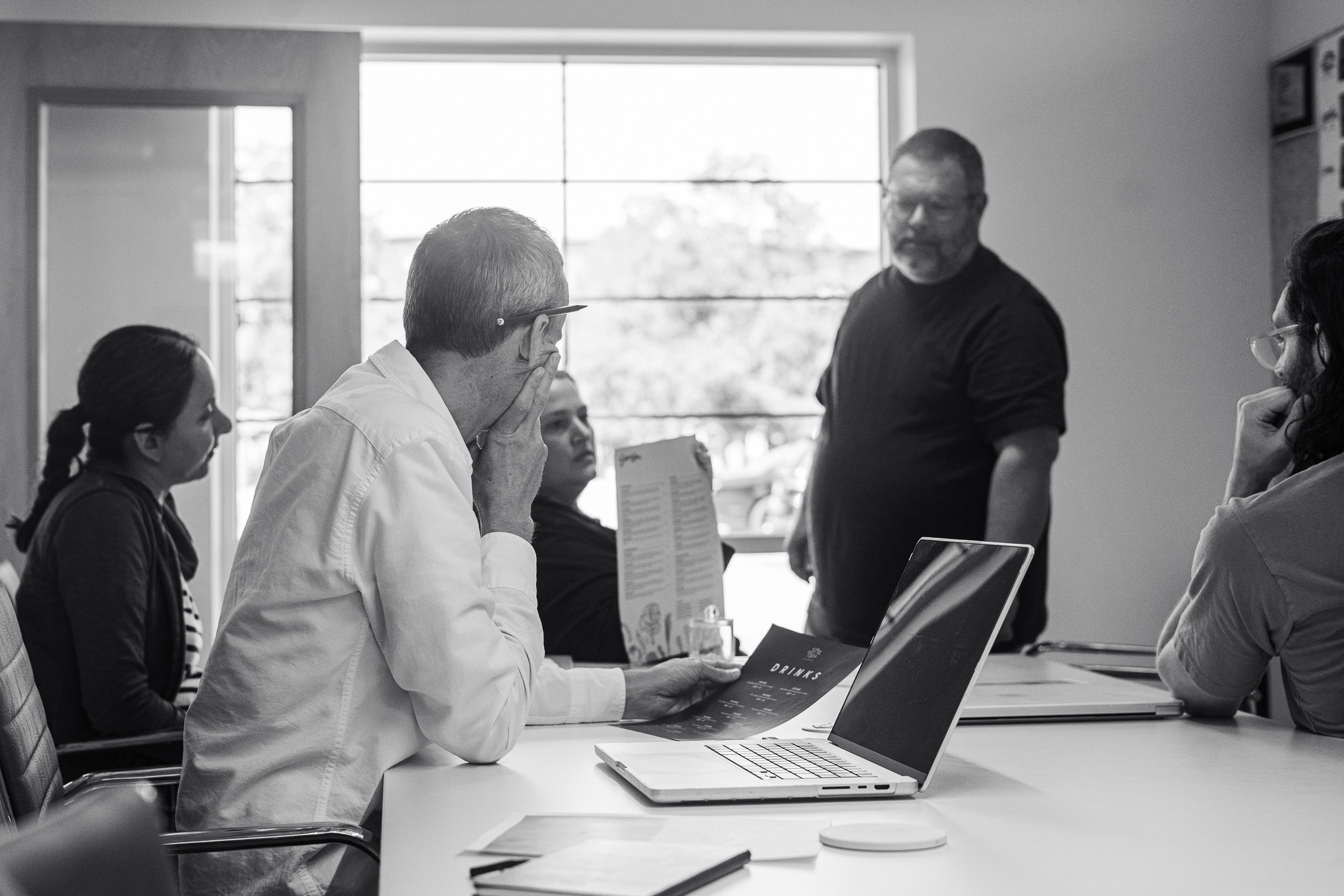 Five people are having a meeting in an office. One man is standing and speaking while the others sit at a table with laptops and papers. The scene is in black and white.