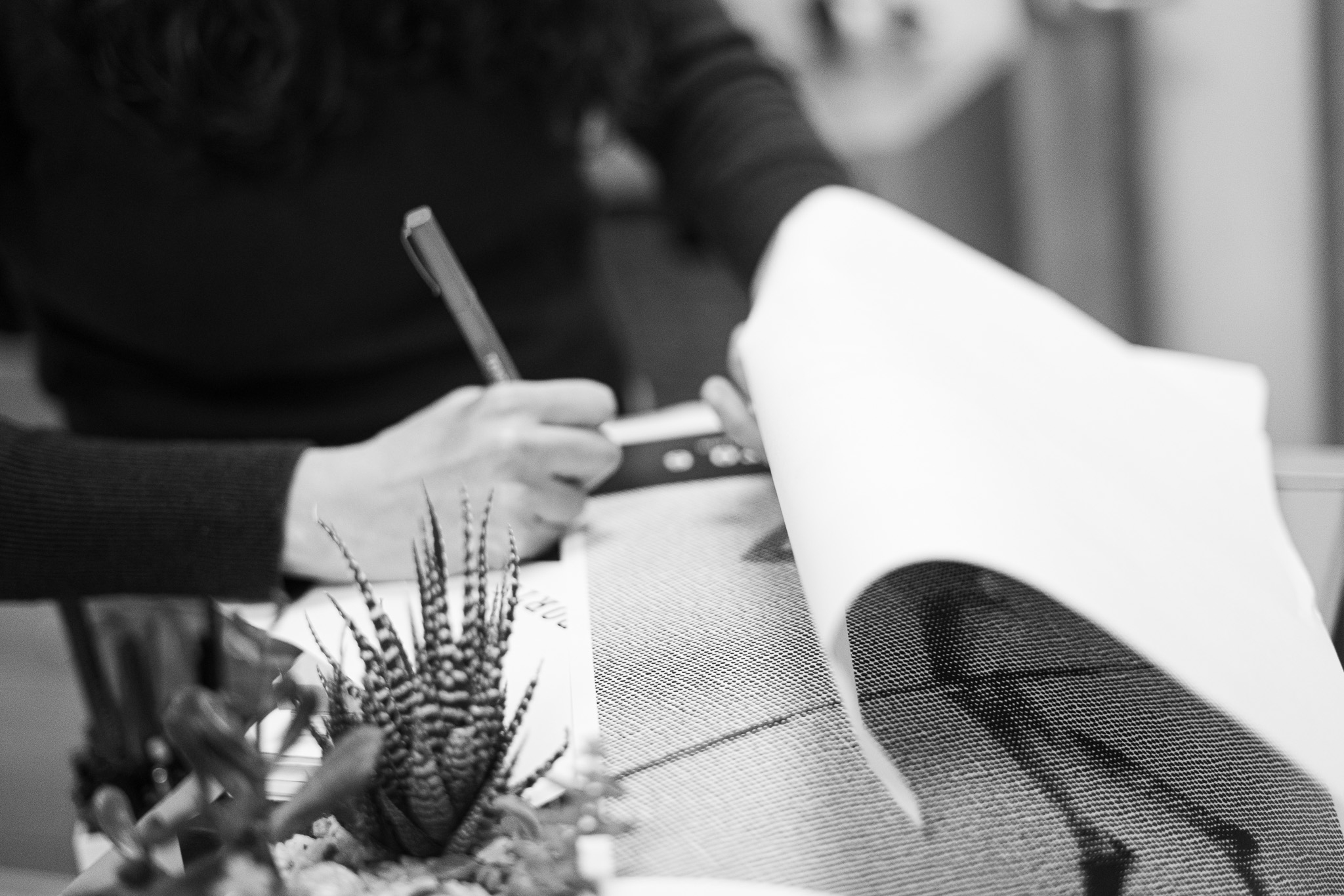 A person writes on a piece of paper at a desk with a potted plant in the foreground; the scene is in black and white.