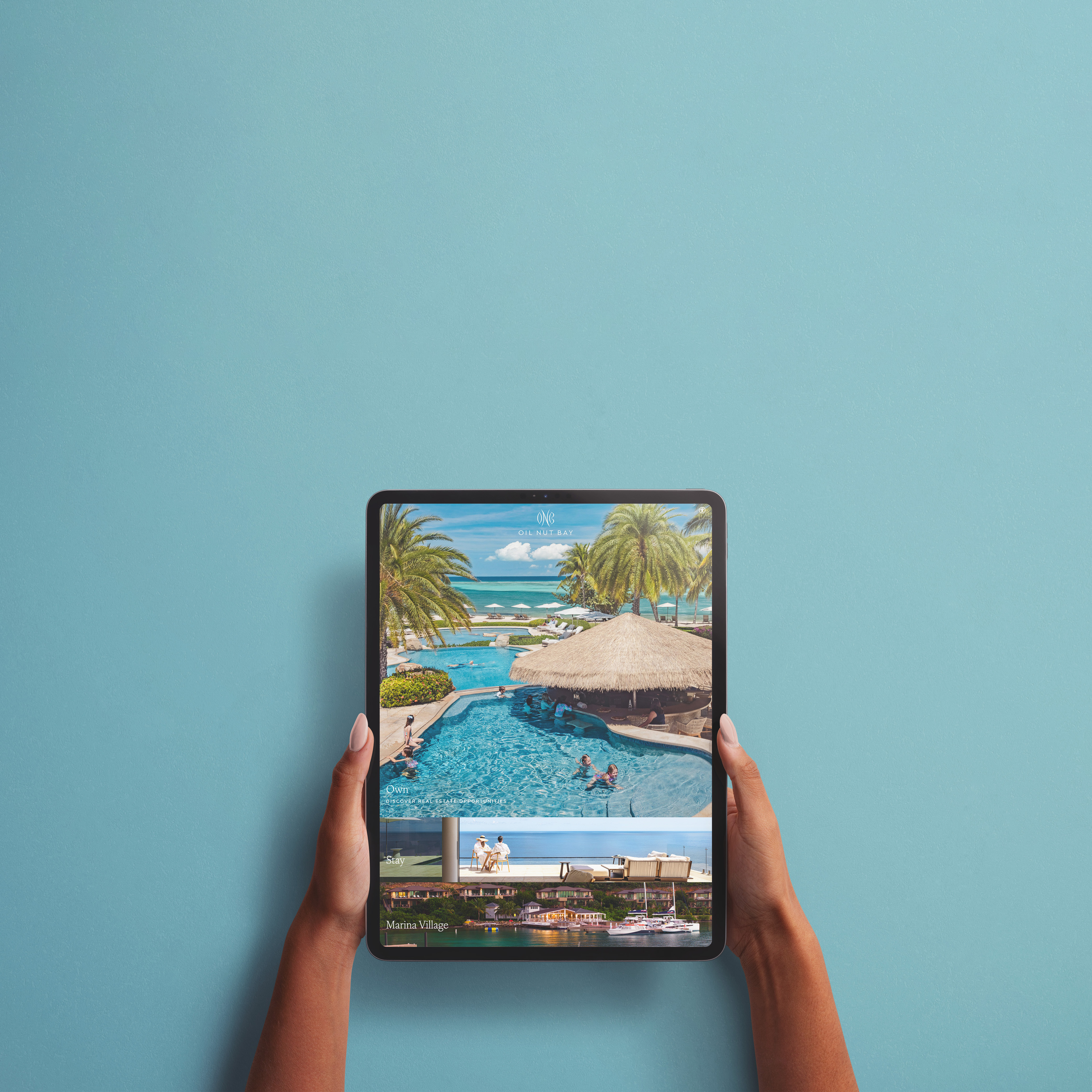 Person holding a tablet displaying a tropical resort scene with a pool, palm trees, thatched-roof hut, and ocean view, against a plain blue background.