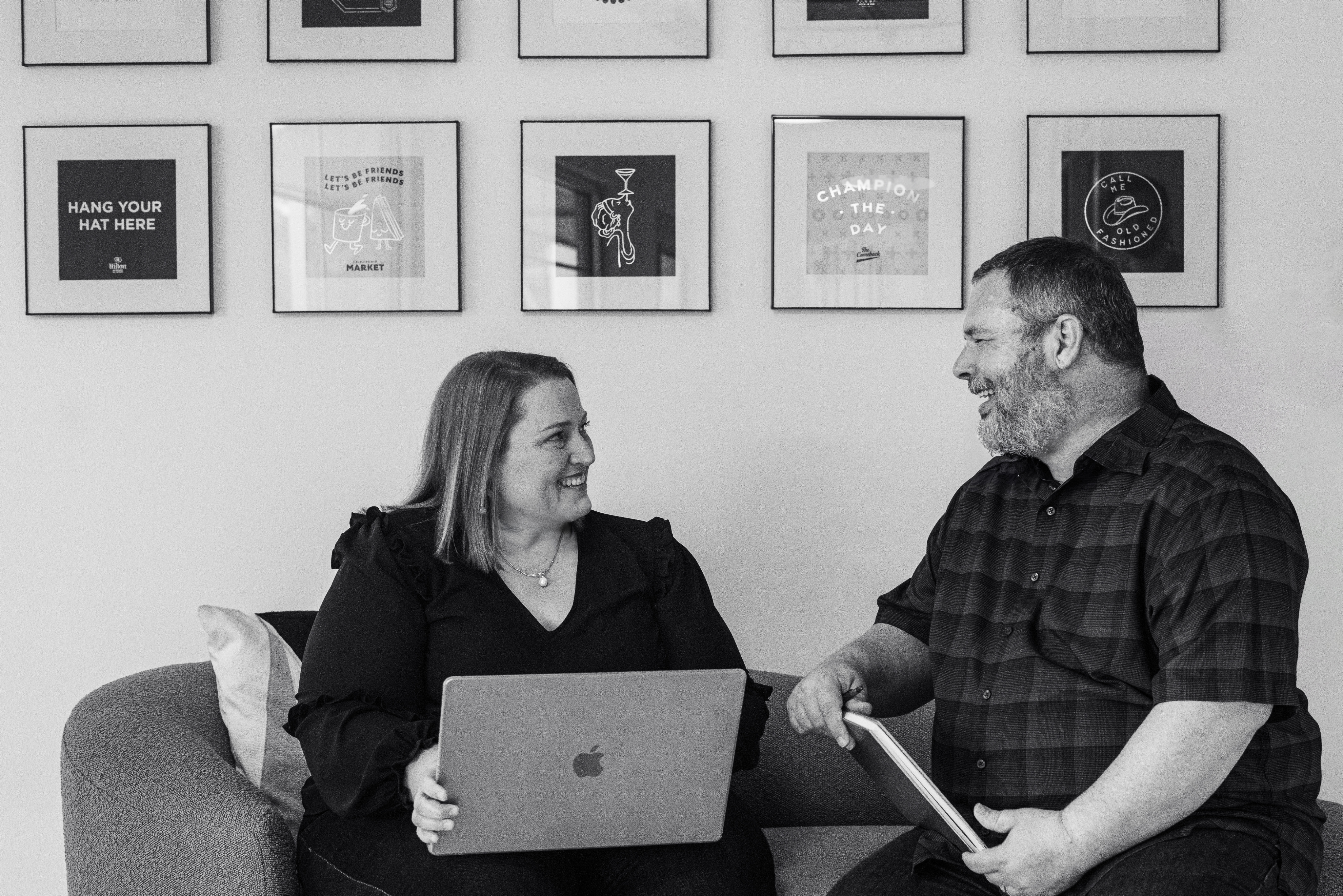 A woman with a laptop and a man with a notebook sit on a couch, smiling and talking; framed pictures are arranged on the wall behind them.
