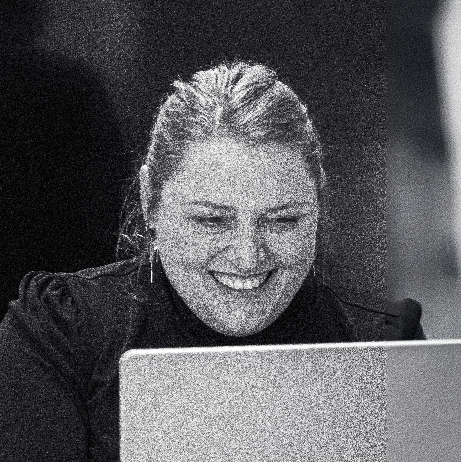 A woman with light hair pulled back smiles while looking at a laptop screen in a dimly lit room.