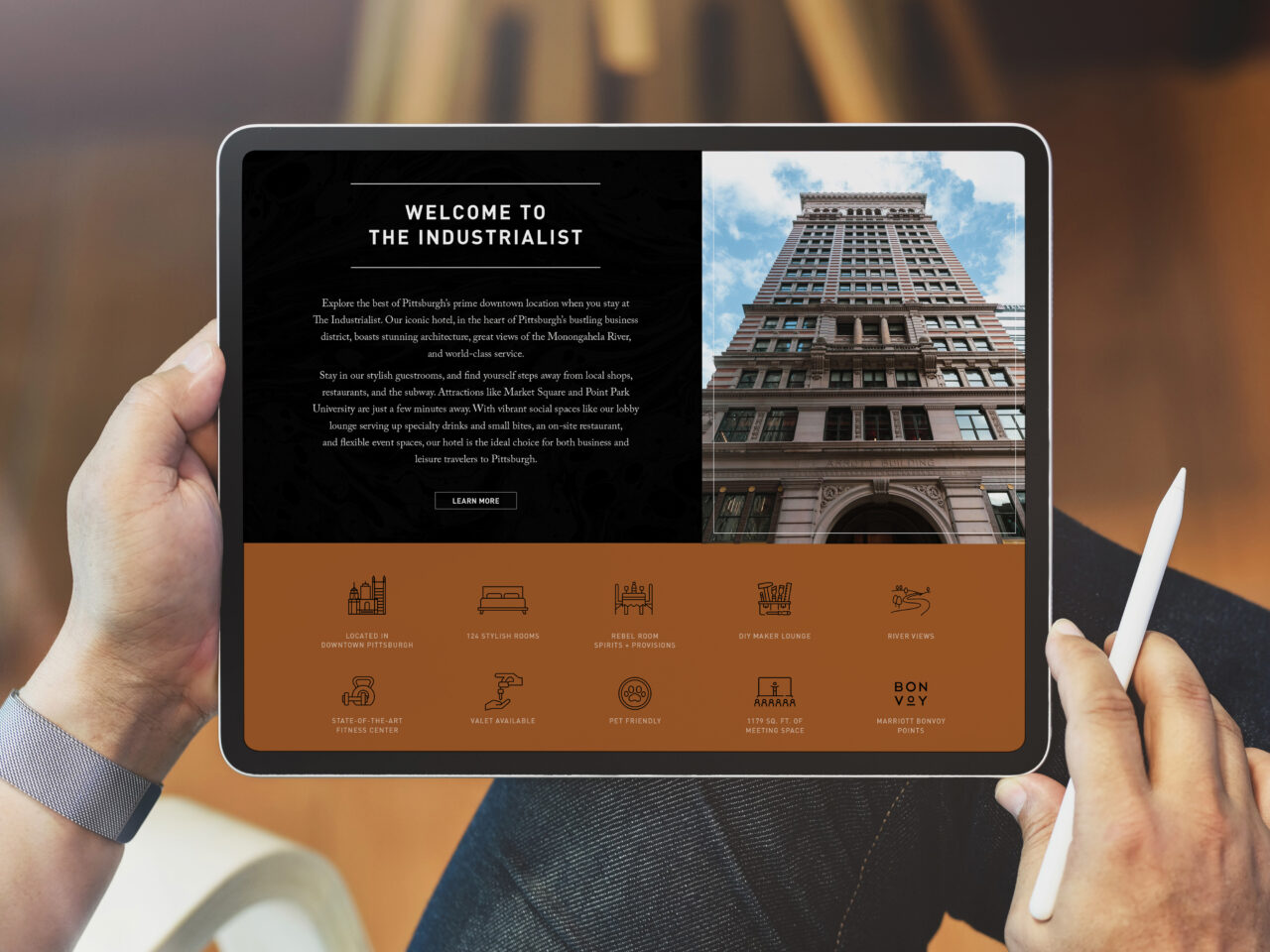 Person holding a tablet displaying a website for The Industrialist hotel, featuring images, descriptive text, and amenity icons with a stylus in hand.