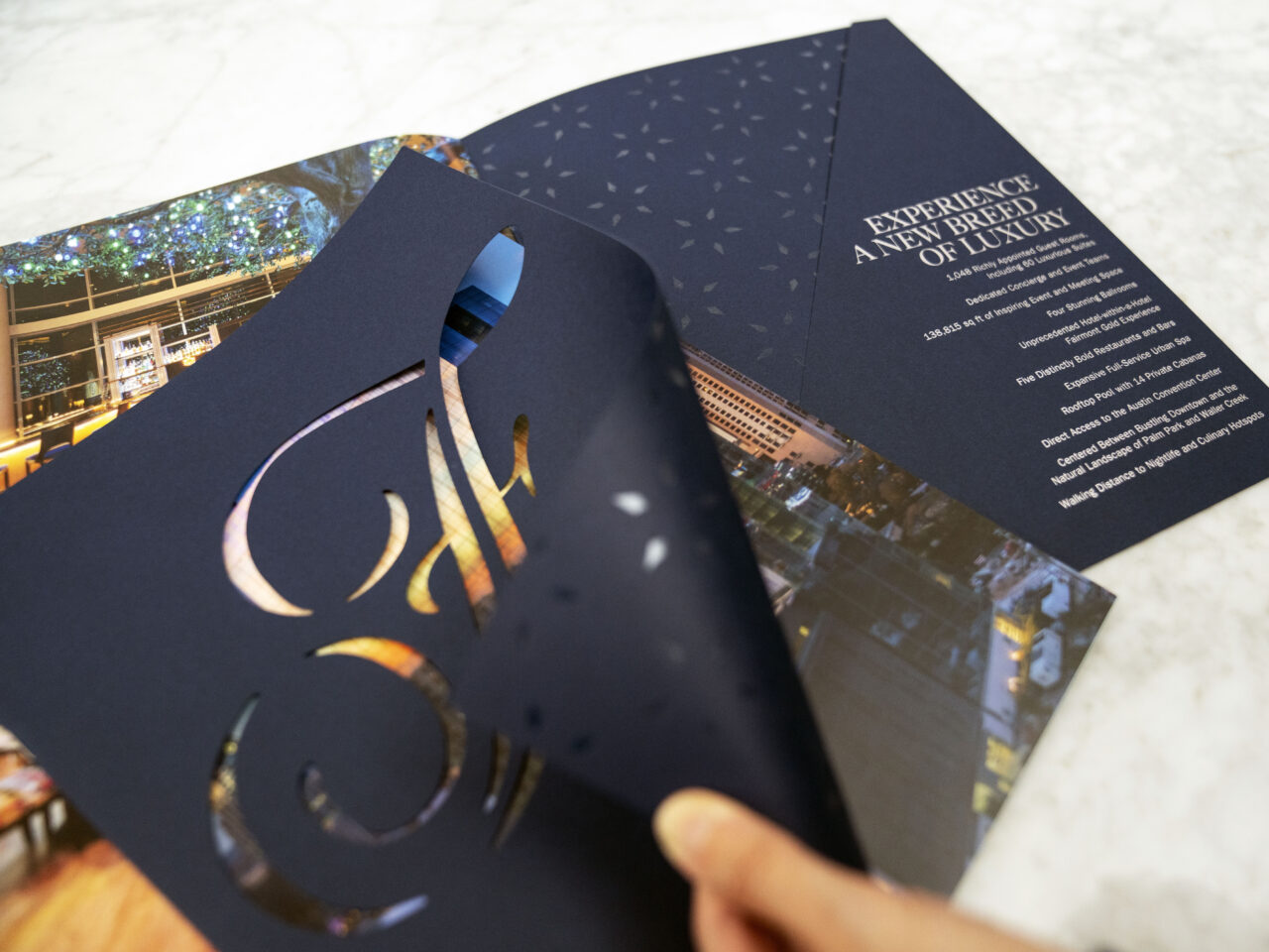A hand lifts a navy blue brochure cover with a cut-out design, revealing a luxury hotel advertisement and text inside.