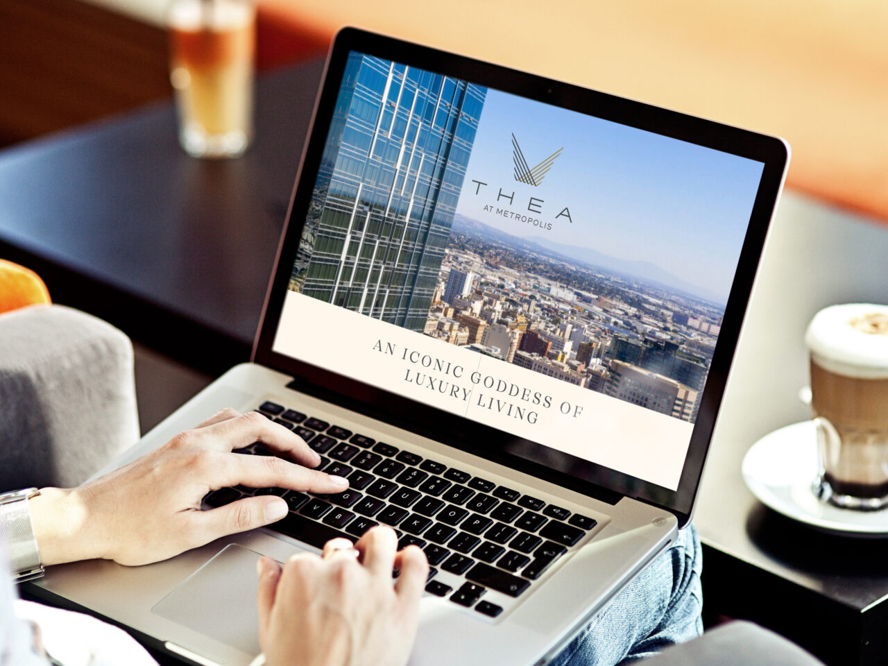 Person using a laptop displaying a website for "THEA at Metropolis," featuring cityscape images and the slogan "An Iconic Goddess of Luxury Living." A coffee cup sits nearby on the table.