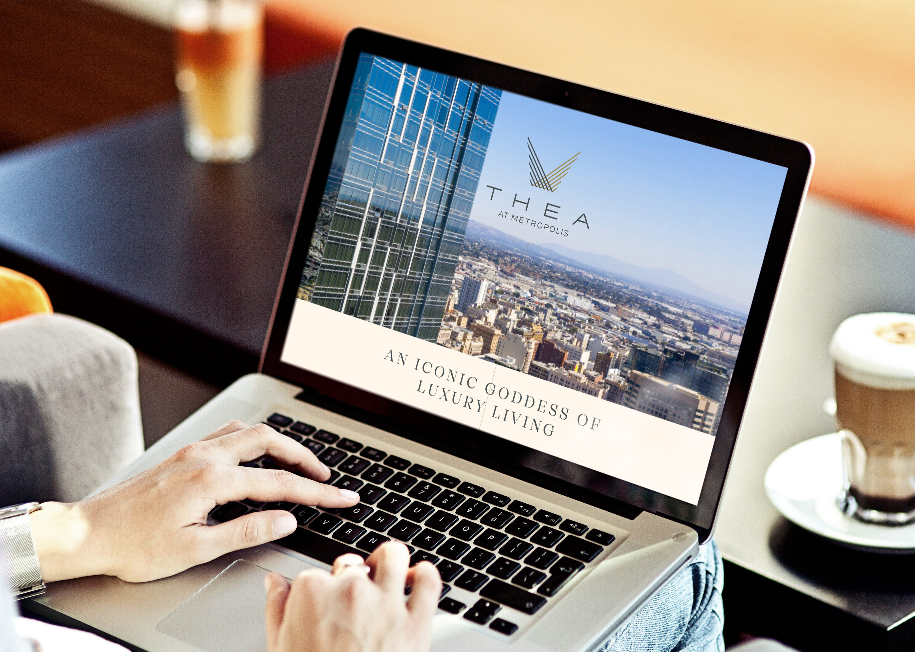 Person using a laptop displaying a website for "THEA at Metropolis," featuring cityscape images and the slogan "An Iconic Goddess of Luxury Living." A coffee cup sits nearby on the table.