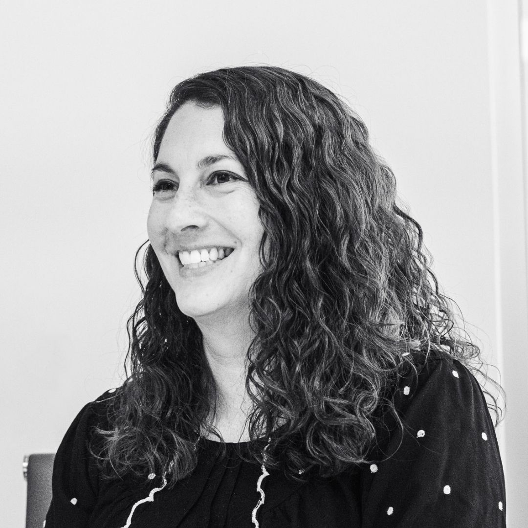 A woman with curly hair and a dark, patterned top is smiling while seated, in a black and white photo against a plain background.