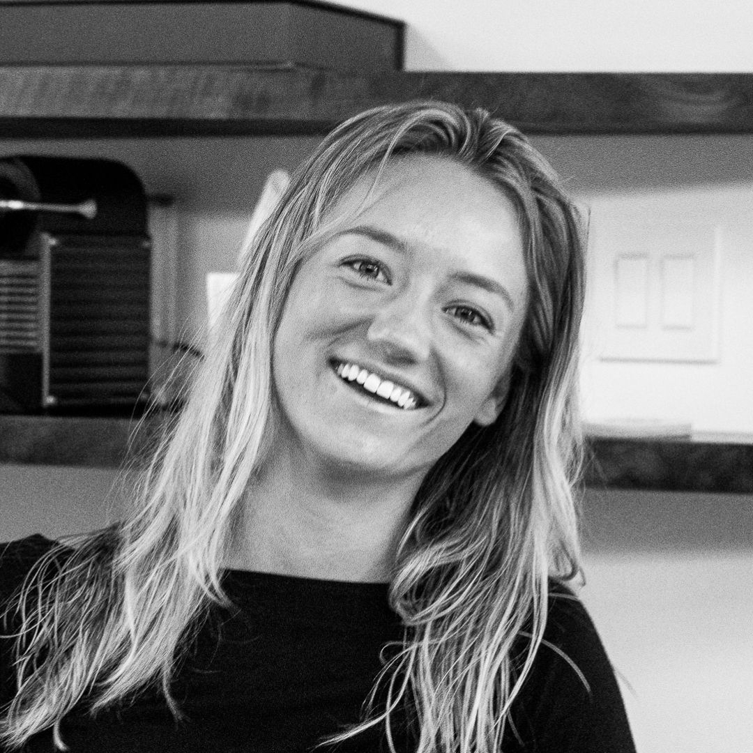 A woman with long, light hair smiles at the camera while standing indoors in front of shelves and a wall switch.