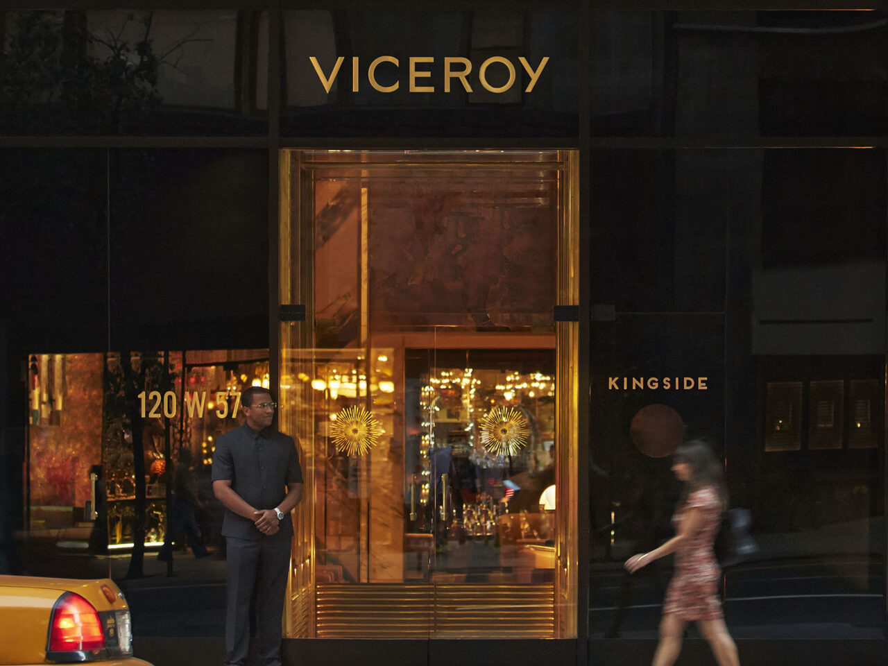 A doorman stands outside the gold-accented entrance of Viceroy hotel, with the Kingside restaurant sign visible and a woman walking past on the sidewalk.