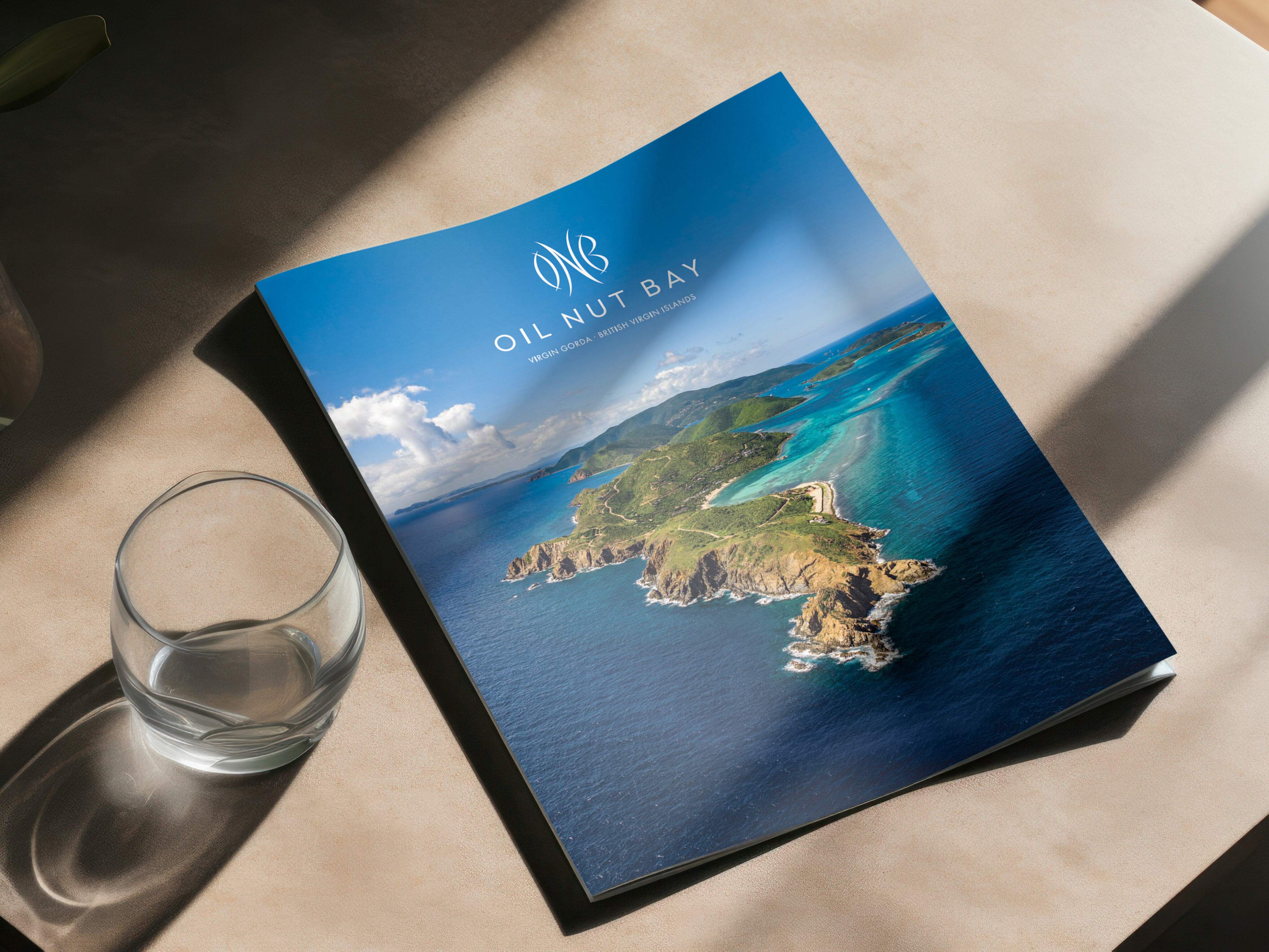 A brochure with an aerial view of coastal land and blue water, titled "Oil Nut Bay," lies on a table next to an empty glass.