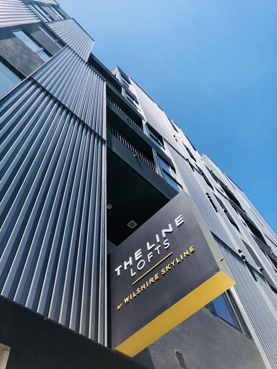 A low-angle view of The Line Lofts building shows its modern facade and a sign reading "THE LINE LOFTS" and "WILSHIRE SKYLINE" against a clear blue sky.