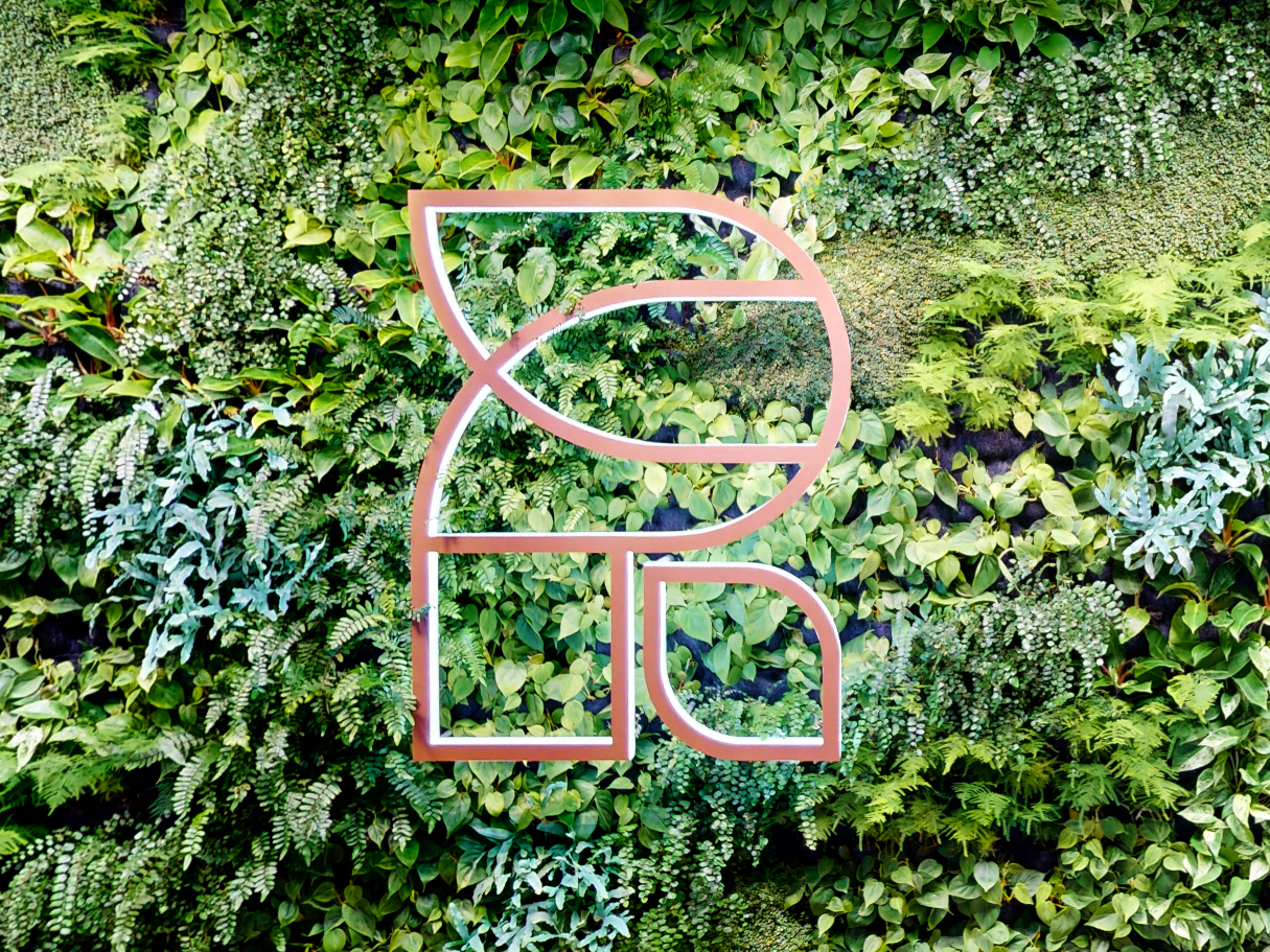 A stylized geometric logo in light pink is displayed on a lush green vertical garden made up of various types of foliage and plants.