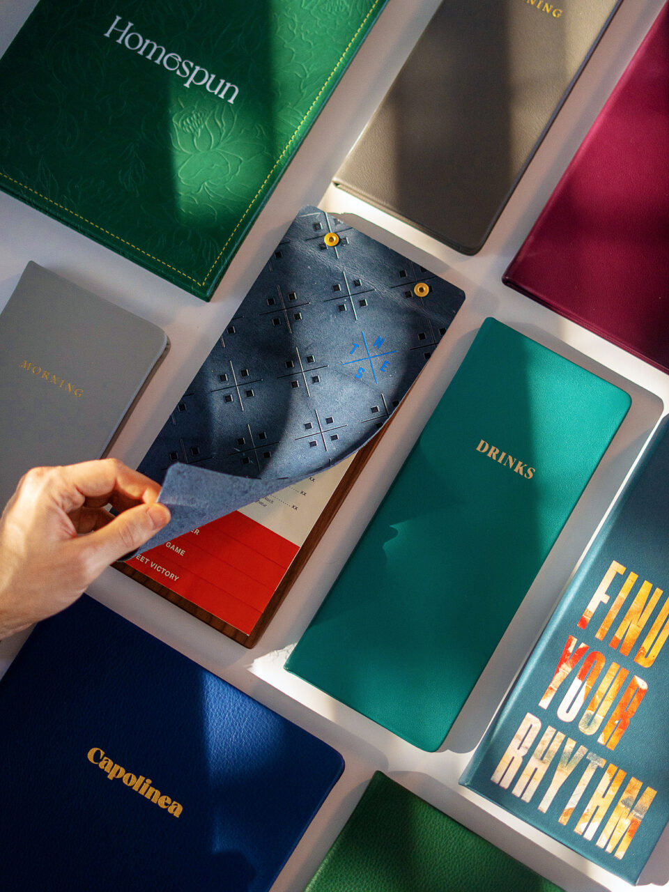 A hand flips through a blue perforated notebook surrounded by various closed, colorful journals and books arranged neatly on a surface.
