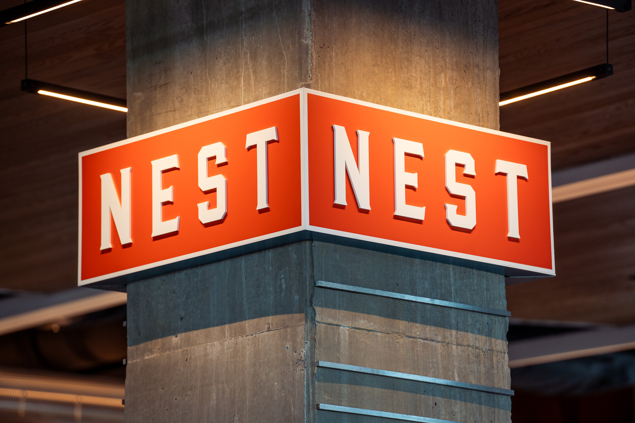 A red rectangular sign with white text reading "NEST" on both visible sides is mounted on a concrete pillar indoors.