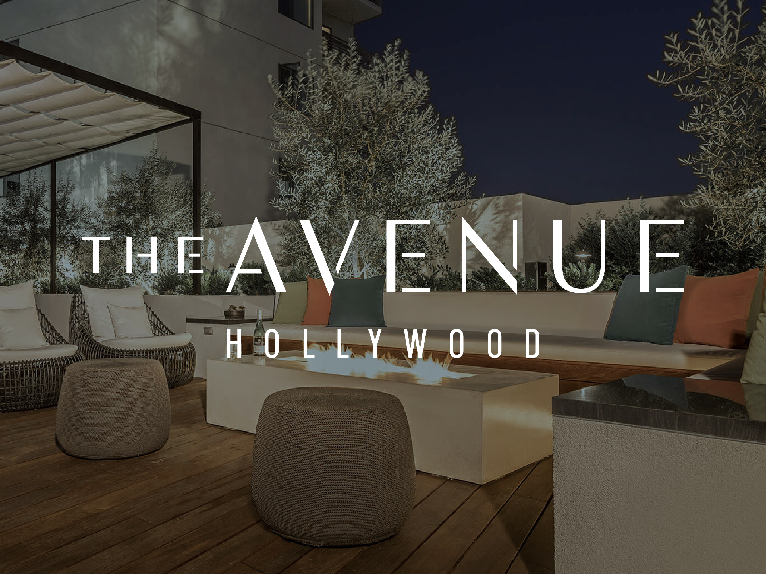 Outdoor lounge area with modern seating, a fire pit, and greenery at night. Large text over image reads "The Avenue Hollywood.