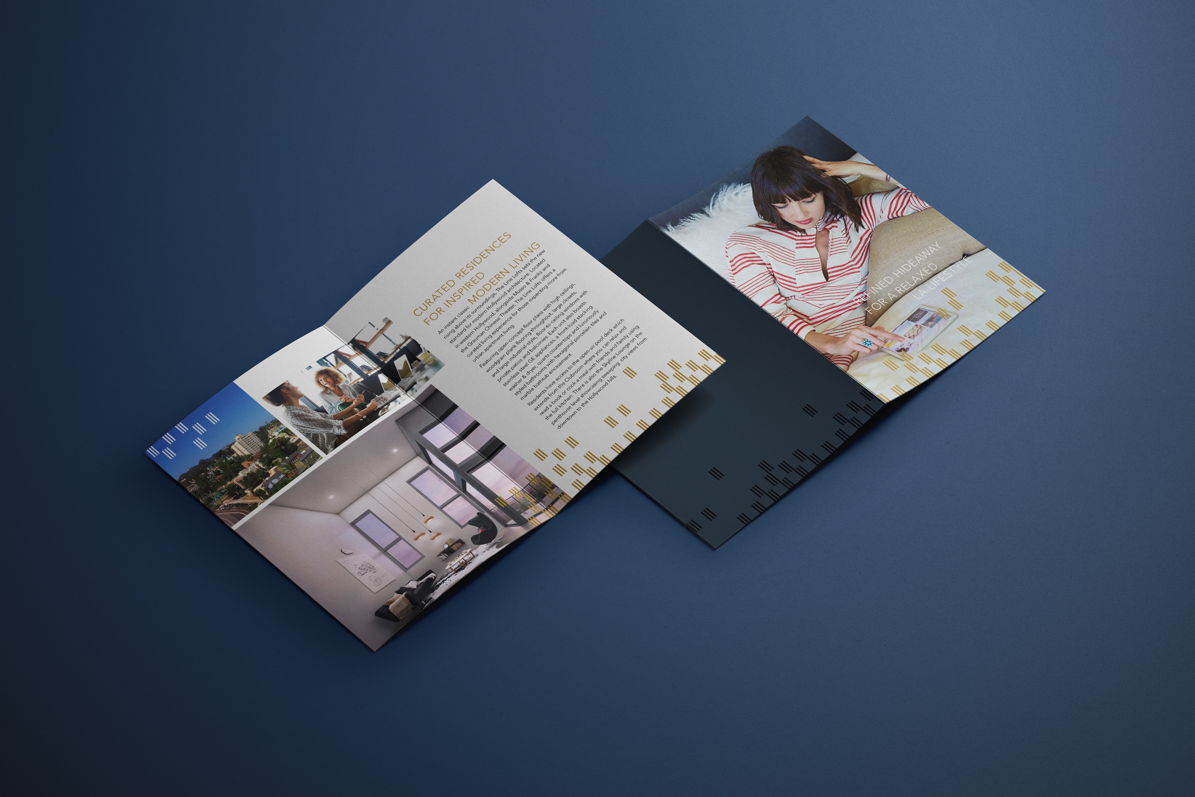 An open brochure on a dark blue surface, showing photos of interiors and a woman reading, with sections of text and decorative gold square patterns.