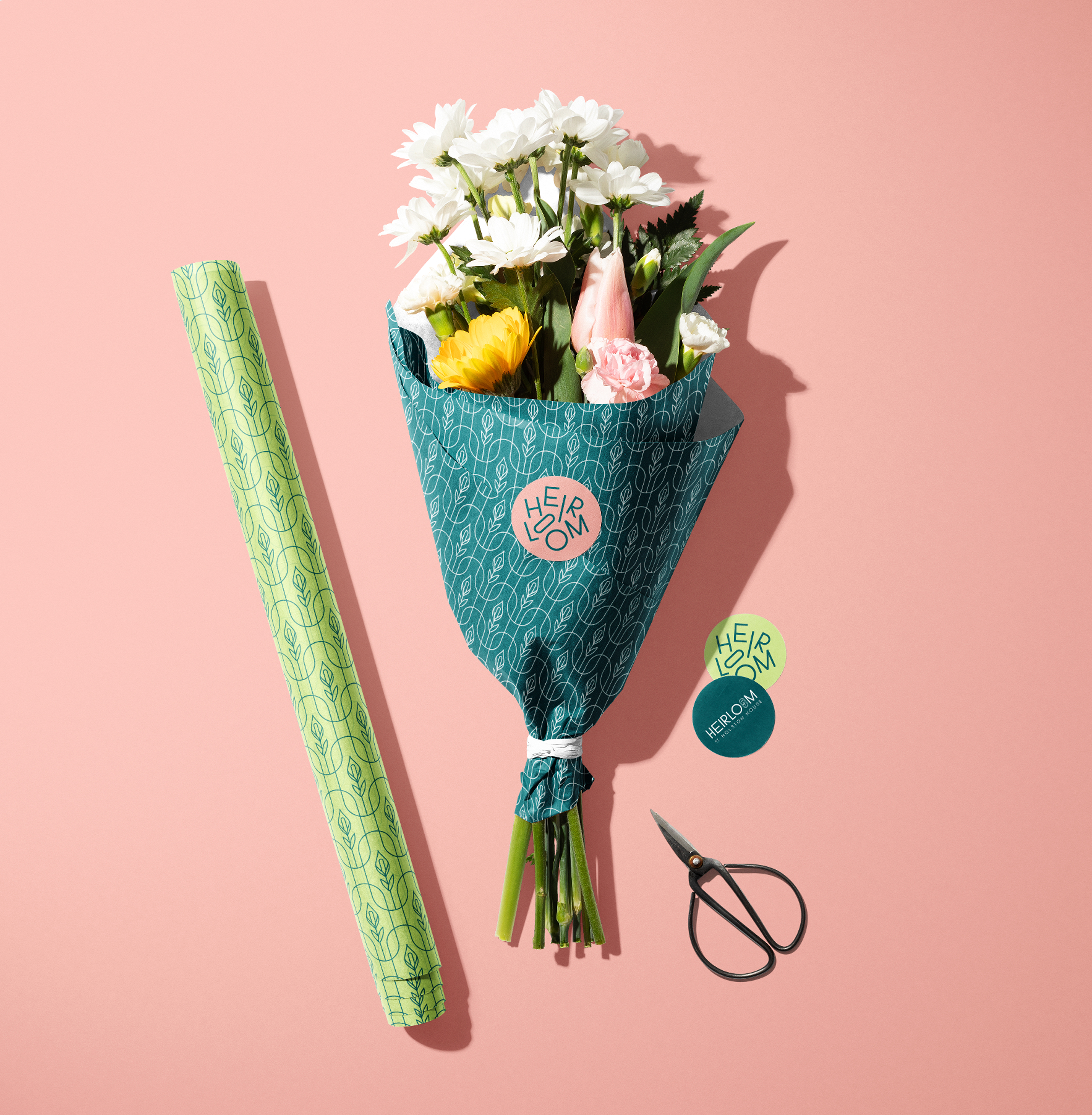 A bouquet of assorted flowers wrapped in blue patterned paper sits on a pink surface next to green wrapping paper, scissors, and two stickers labeled "HEIRLOOM.