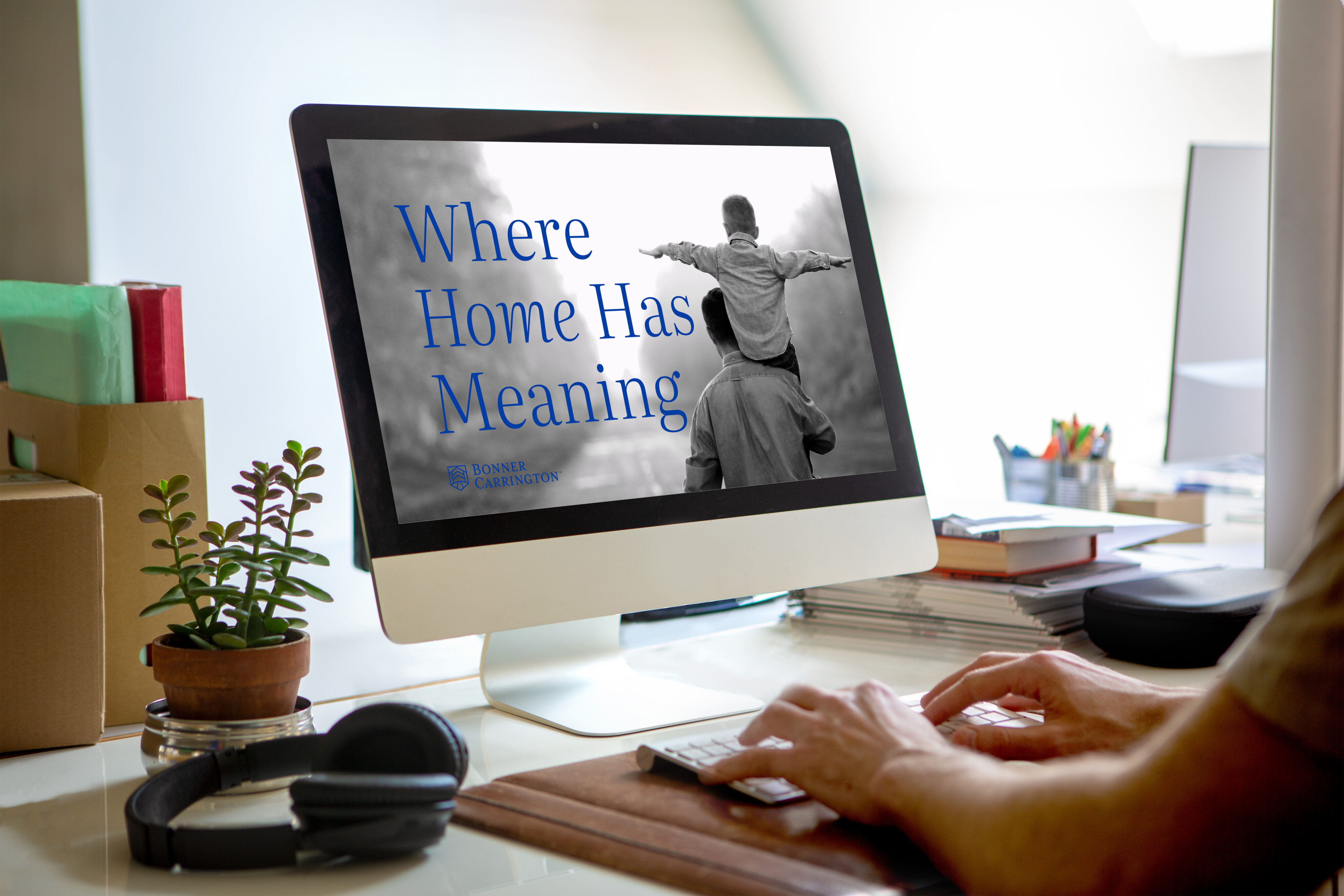 A person types at a desk with a computer screen displaying the text "Where Home Has Meaning" and an image of a child on an adult's shoulders.