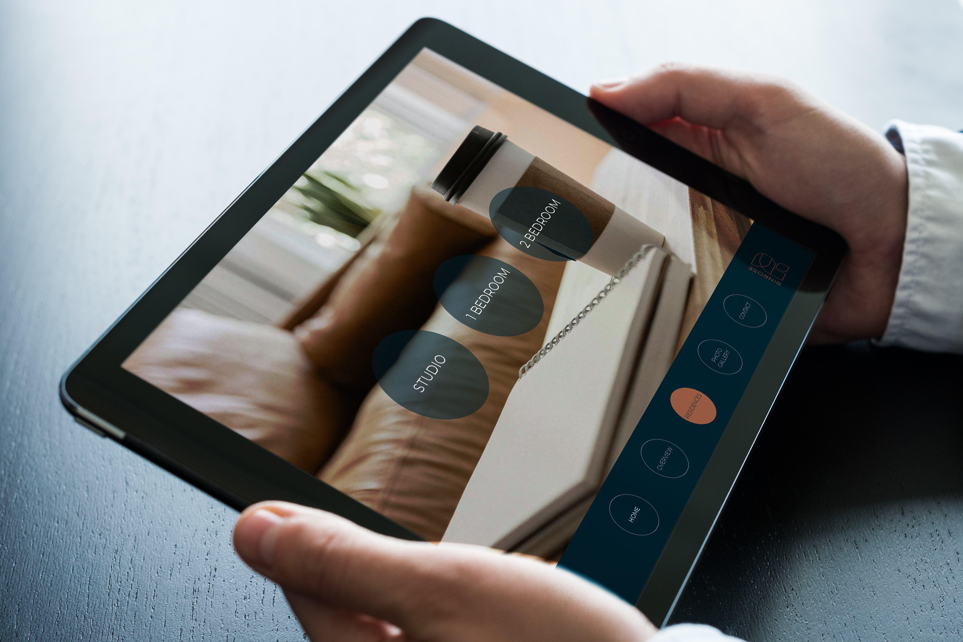Person holding a tablet displaying a home control interface with options for studio, bedroom, and other rooms, against a background showing a couch and window.