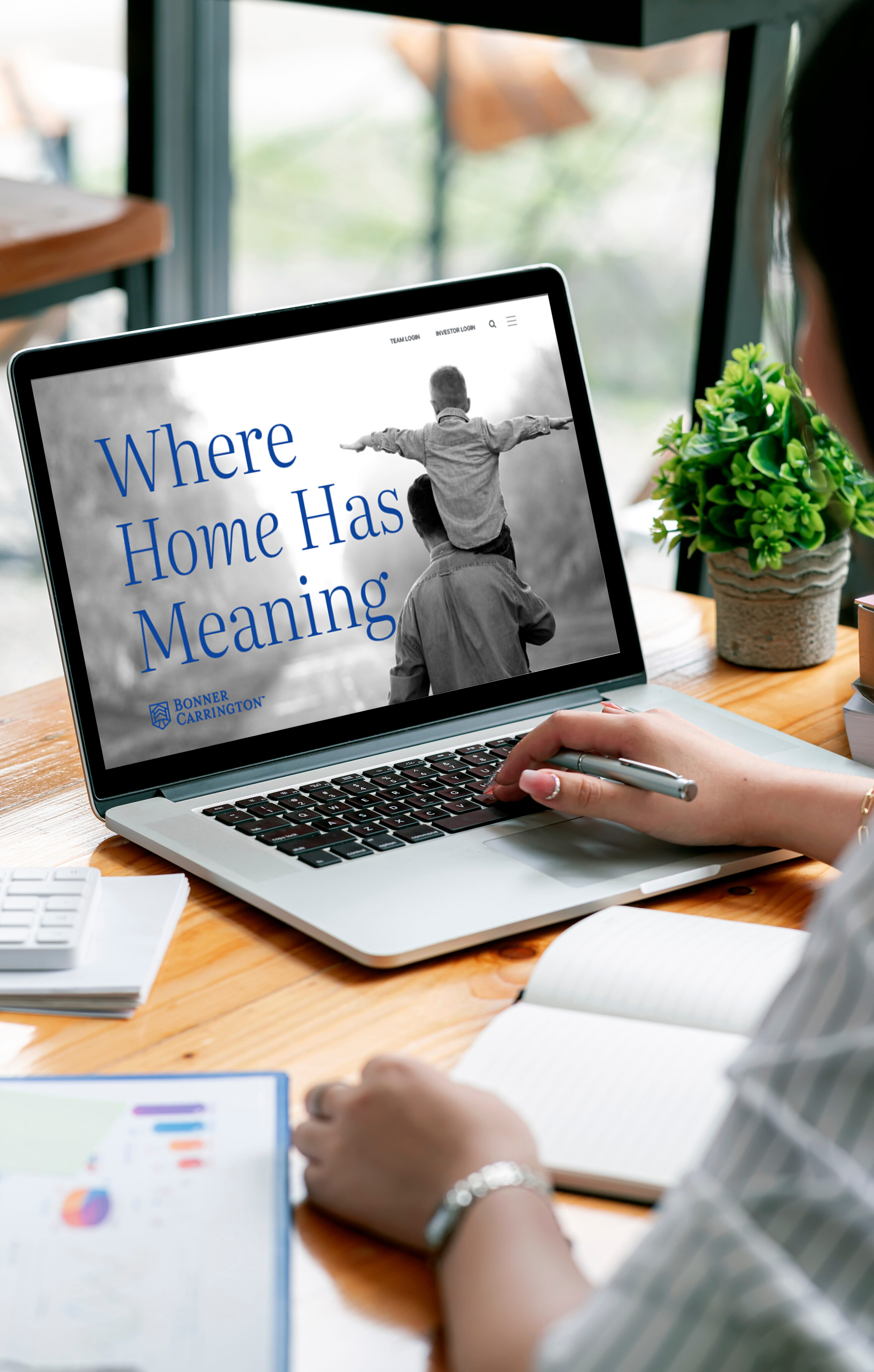 Person working at a desk with a laptop displaying a website that says "Where Home Has Meaning" next to a black-and-white image of an adult carrying a child on their shoulders.