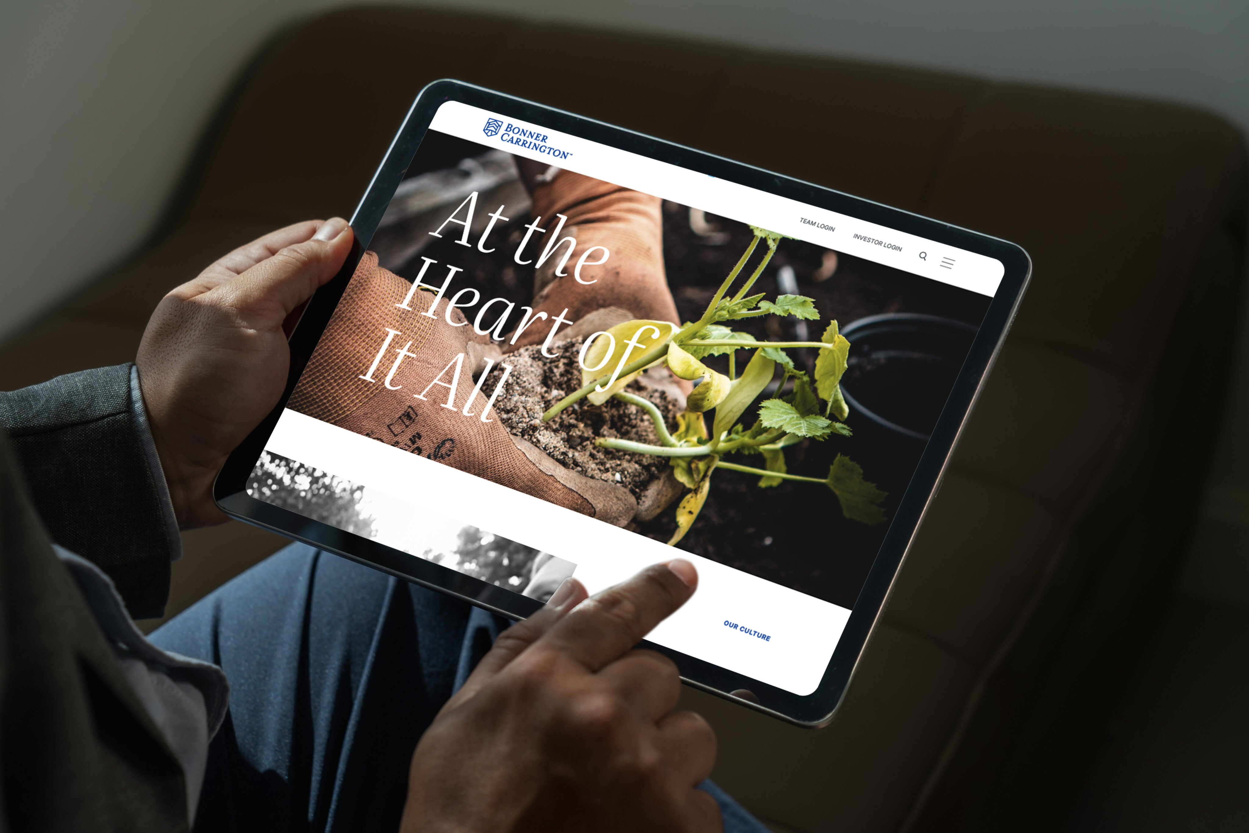 A person holds a tablet displaying a website with an image of hands planting a seedling and the text "At the Heart of It All" on the screen.