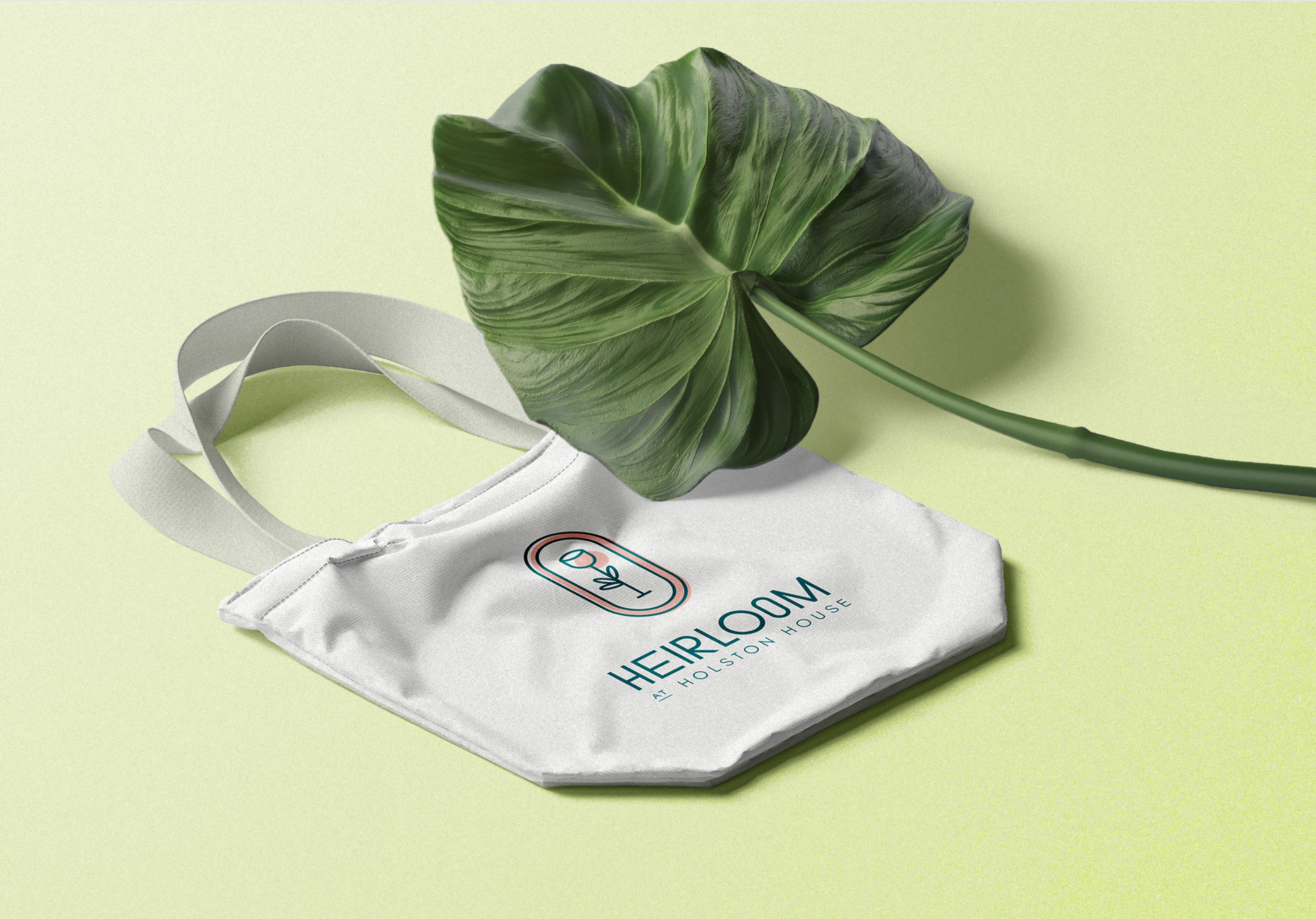 White tote bag with "Heirloom: A Poison House" logo beside a large green leaf on a light green background.