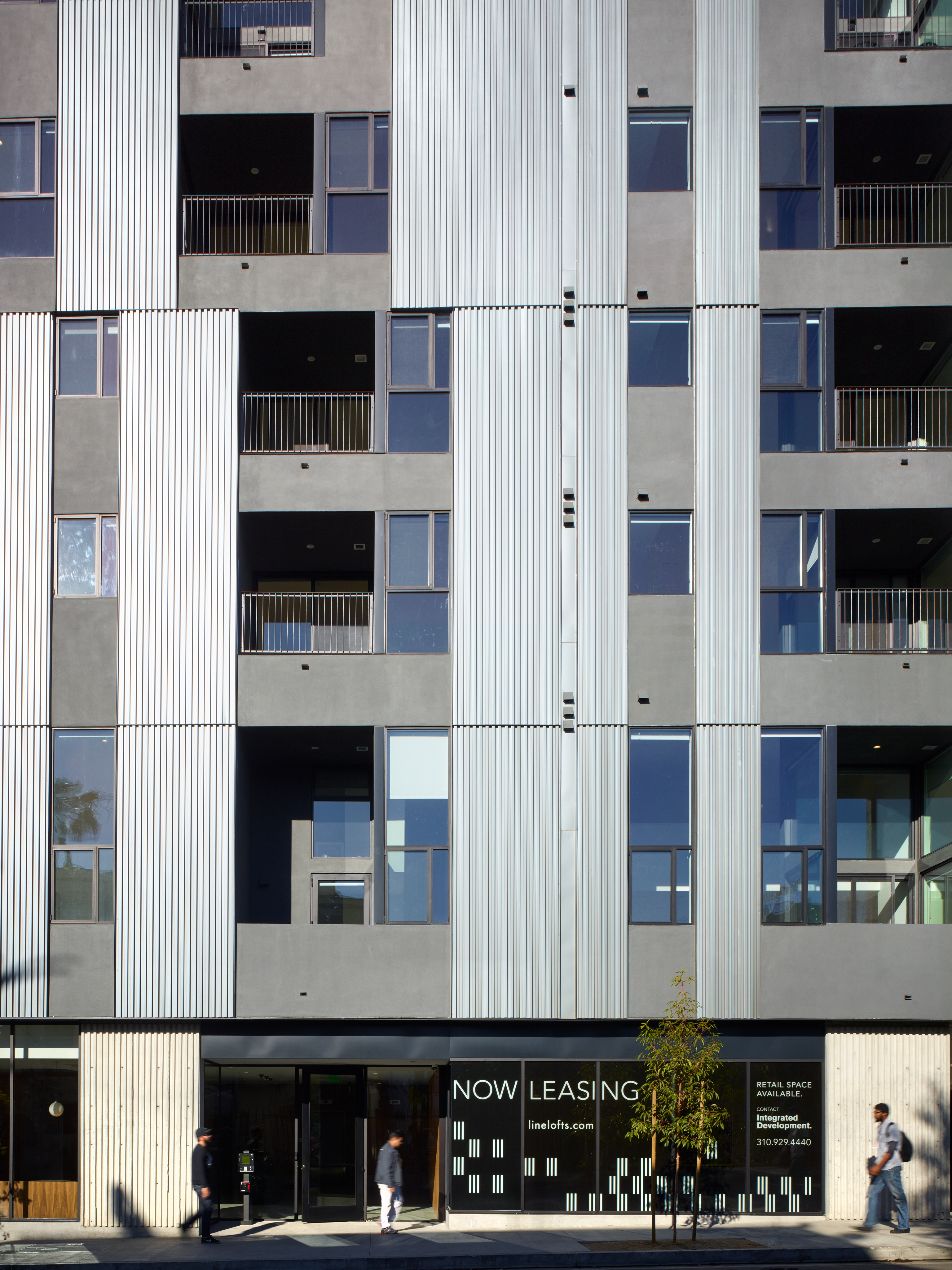 Modern apartment building with gray corrugated metal and concrete exterior; "Now Leasing" sign displayed at street level; several people walking on the sidewalk.