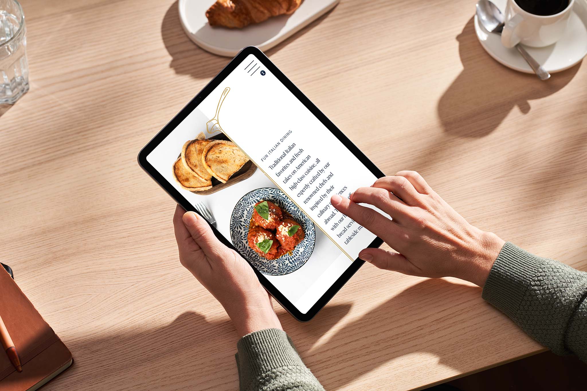 Person holding a tablet displaying a recipe with a photo of toast and a dish; coffee, croissant, and glass of water are on a light wooden table.