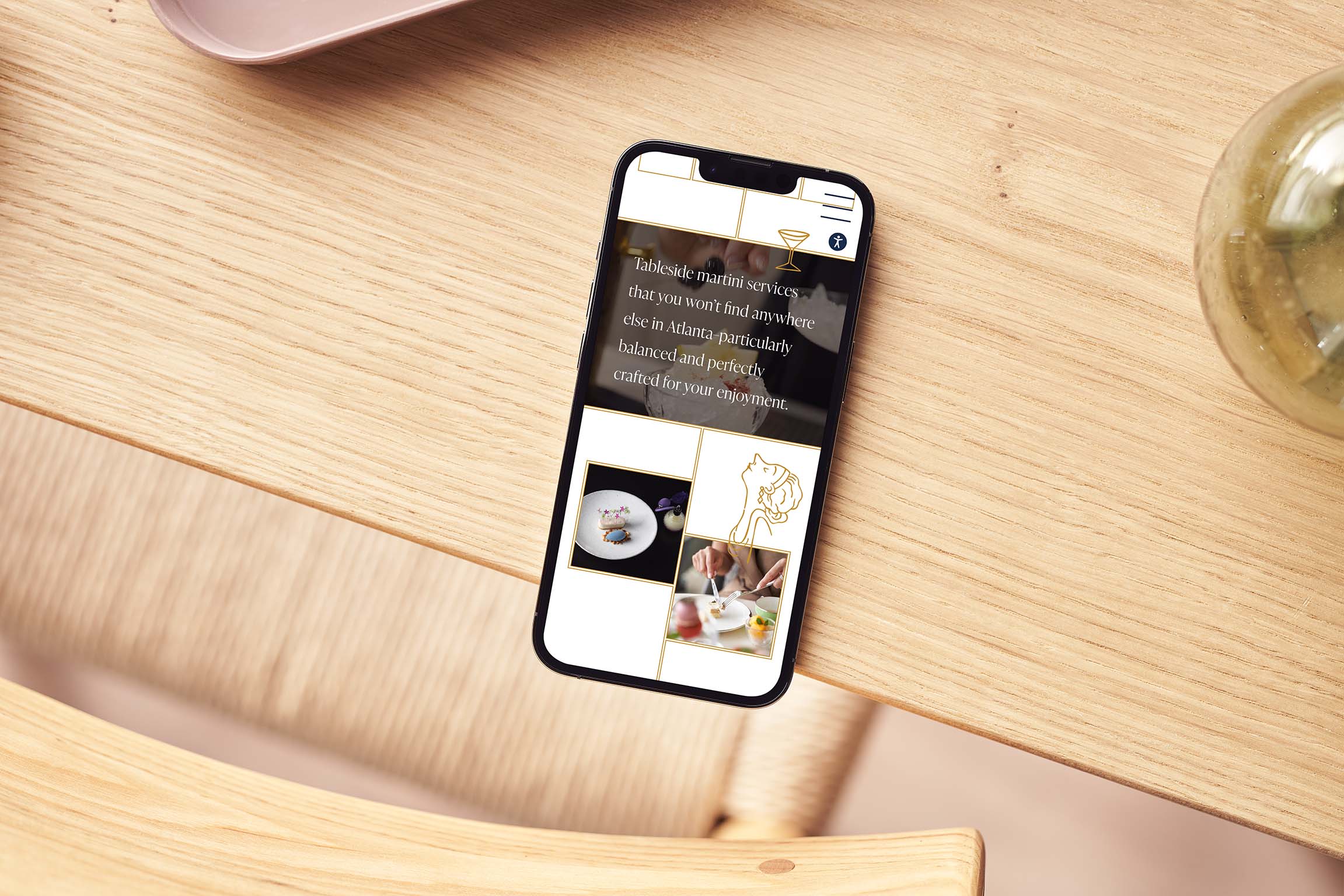 A smartphone displaying a webpage with images and text rests on a light wooden table next to a glass vase and a wooden chair.