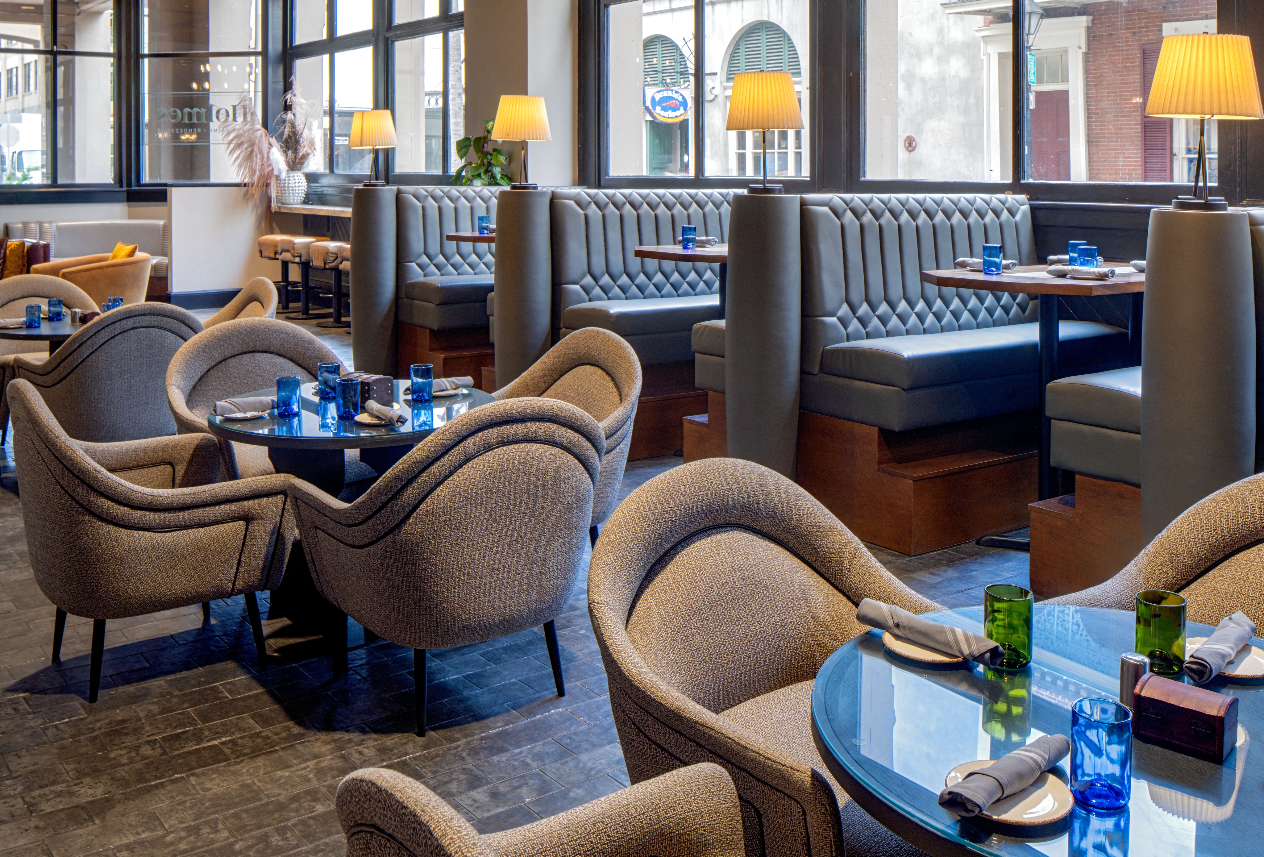 Modern restaurant interior with round tables, cushioned chairs, blue glassware, and booth seating near large windows. Table settings include napkins and glass cups.