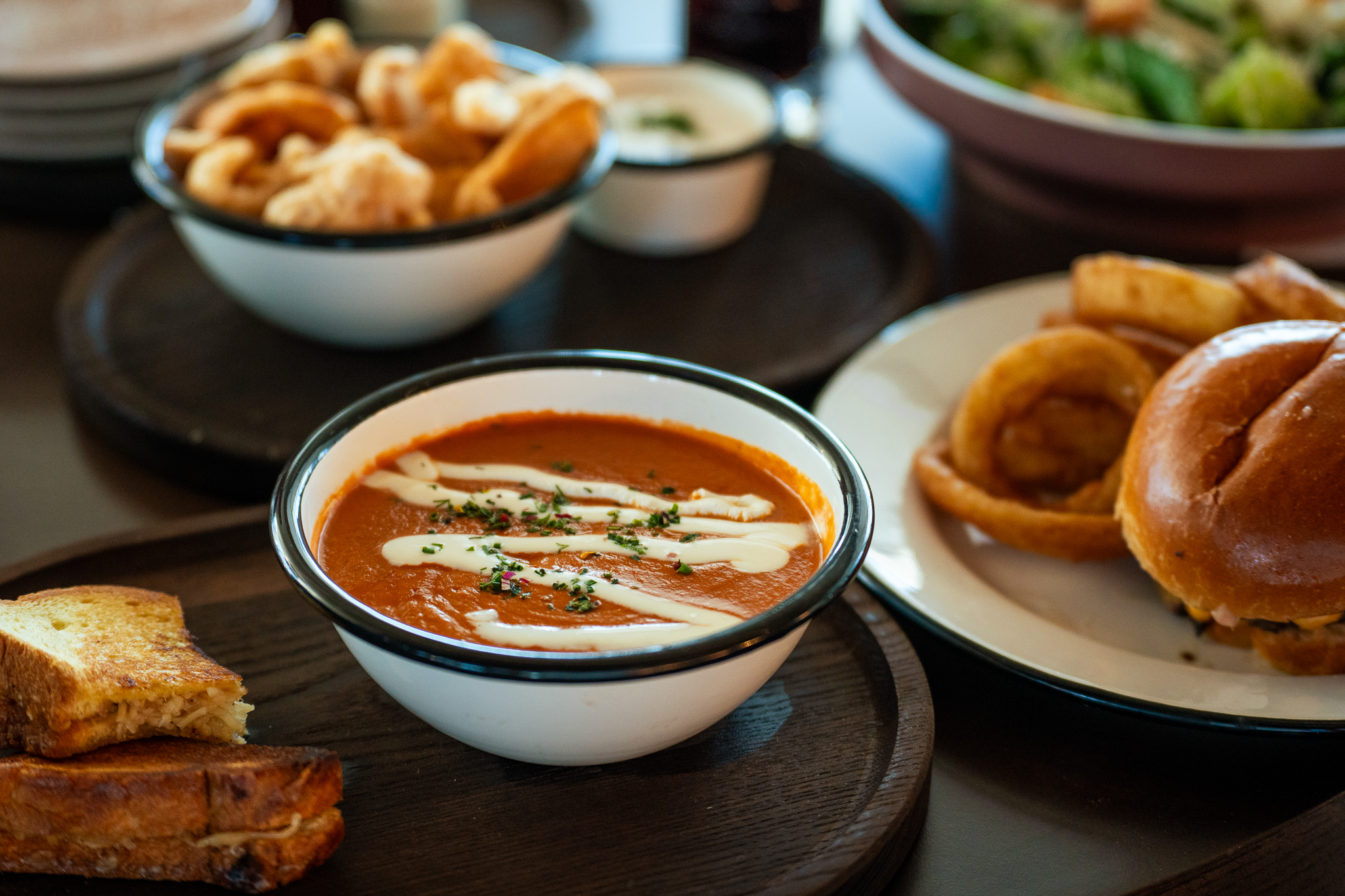 A bowl of tomato soup topped with cream and herbs sits on a wooden tray beside grilled bread, with onion rings, a sandwich, and salad in the background.