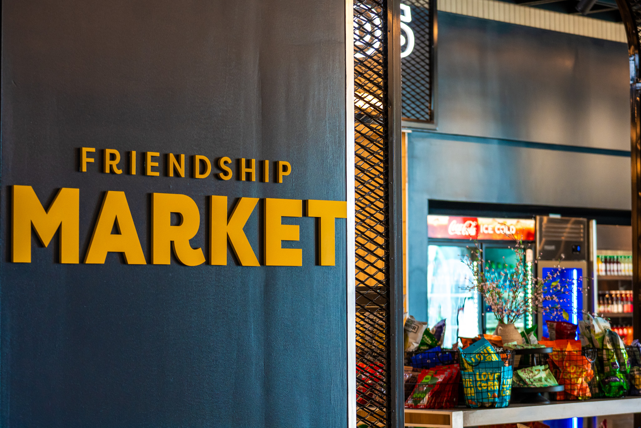 A sign reading "Friendship Market" is displayed on a wall; in the background, snacks and refrigerated drinks are visible inside the market.